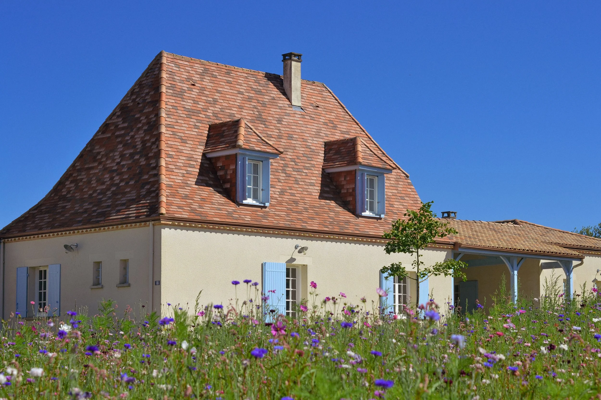 A white house with a steep, brown roof and a porch, surrounded by colorful flowers in pink, white, purple, and blue, with a tree on the right and a clear blue sky in the background.