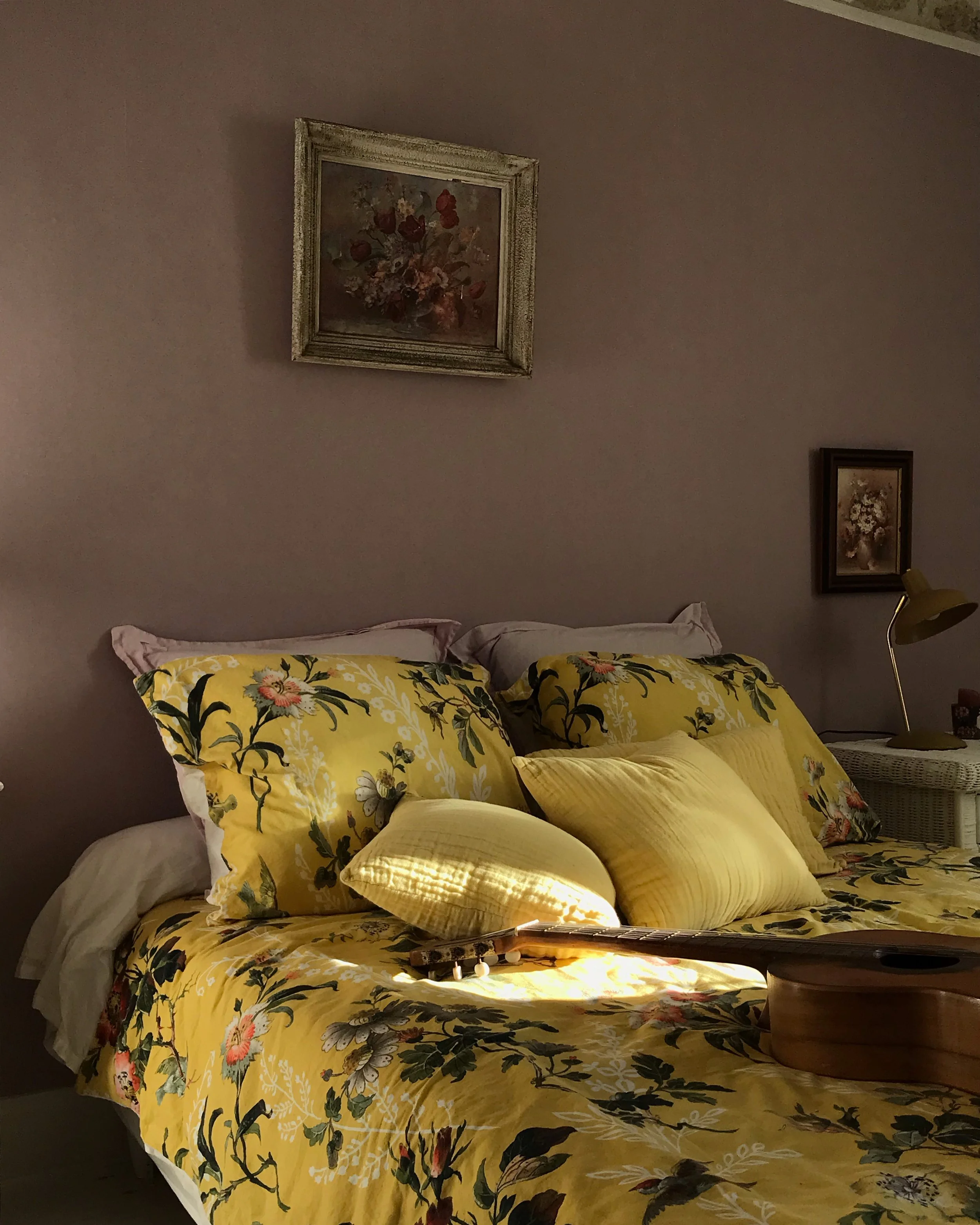 A neatly made bed with white and beige bedding, three decorative pillows, a white nightstand with a lamp, clock, and pink flowers, a window with natural light, and a wall mirror with a floral design above the bed.
