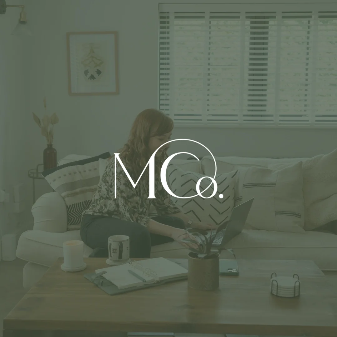 A woman sitting on a sofa with a laptop on her lap, surrounded by various objects on a wooden coffee table, in a cozy living room with a large window and modern decor.