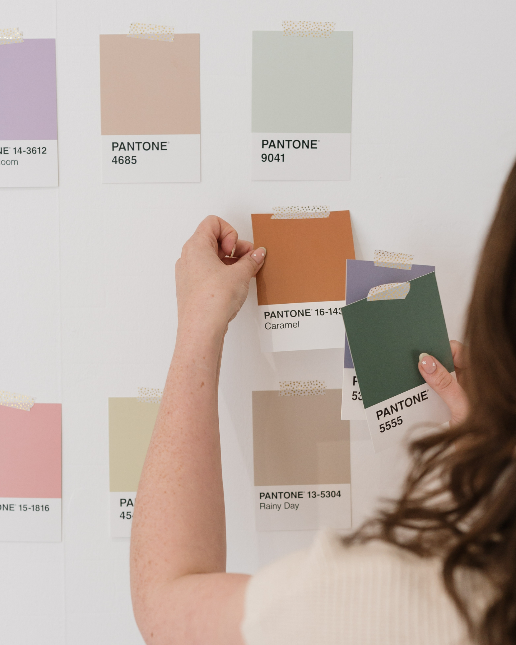 Sammi of Make it Pretty holding colour swatches with Pantone colour codes, with some colour samples pinned on a white wall. UK Brand Identity Design & Presentation Design