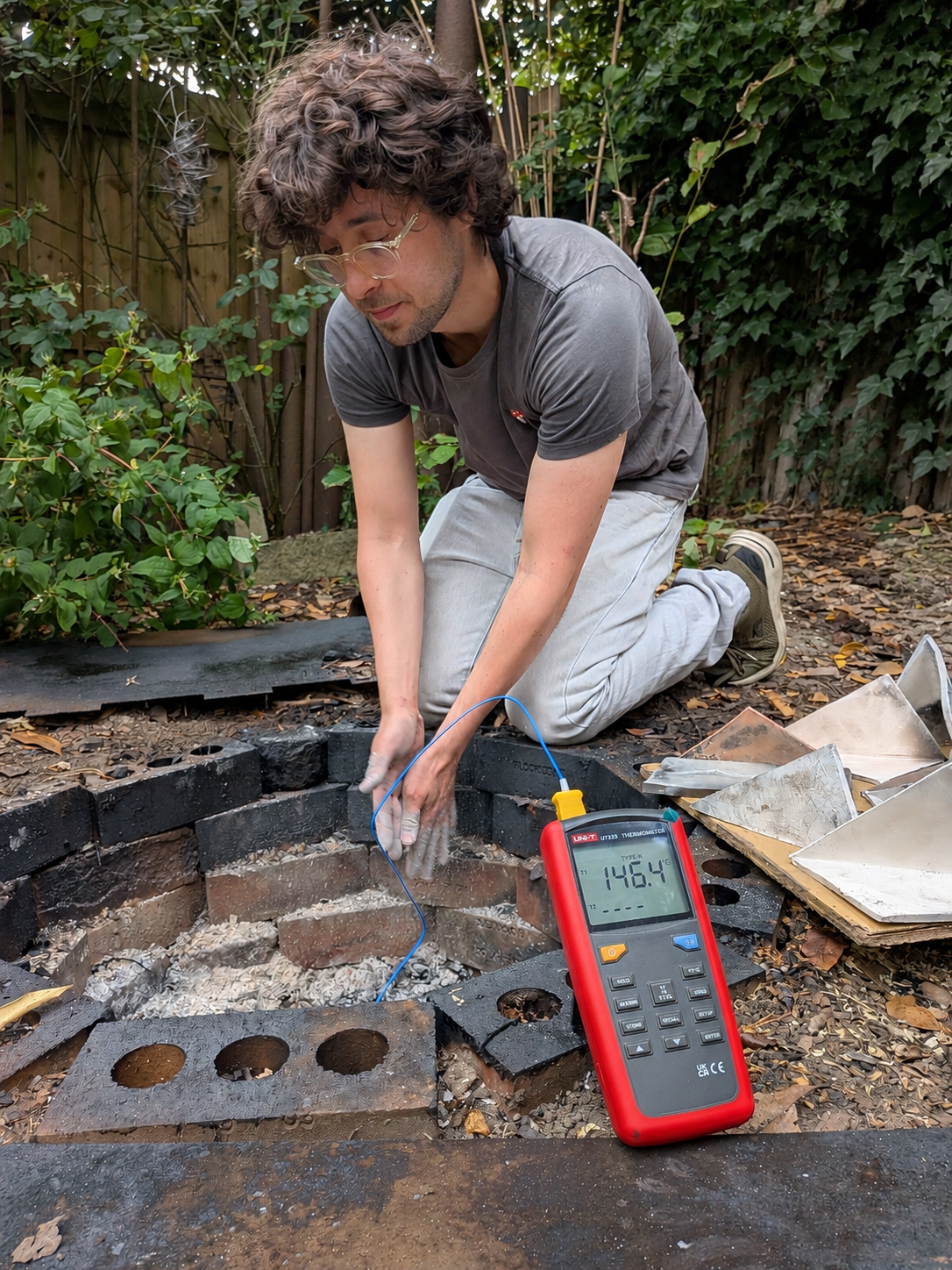 Jacob Chandler white man with curly hair and glasses reaches into brick lined ash filled pit where he is removing pieces from a pit firing.