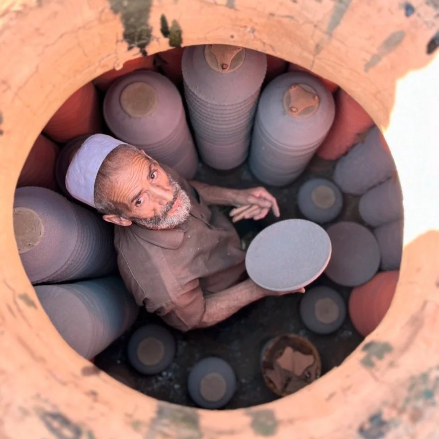 An elderly man with a white cap sitting inside a large clay or ceramic vessel, surrounded by similar vessels, looking up at the camera holding a round object.
