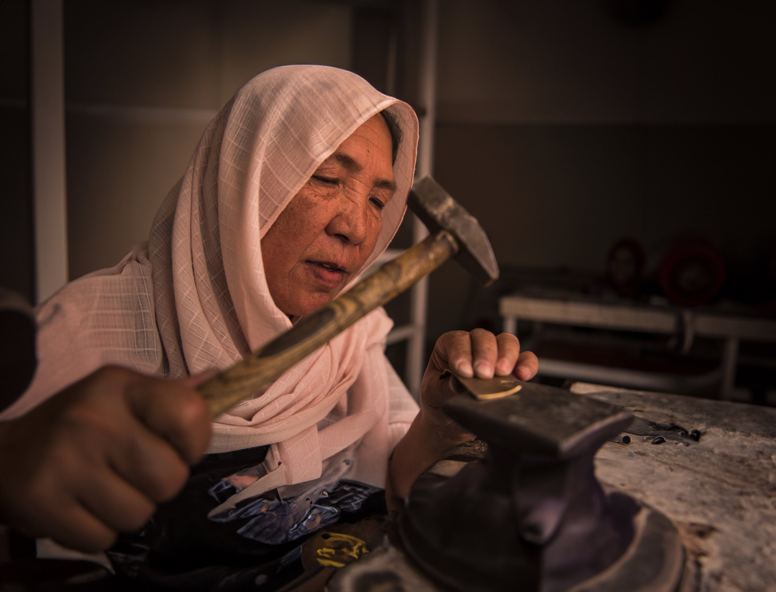 An elderly person with a pink headscarf working with a hammer and chisel on metal in a workshop.