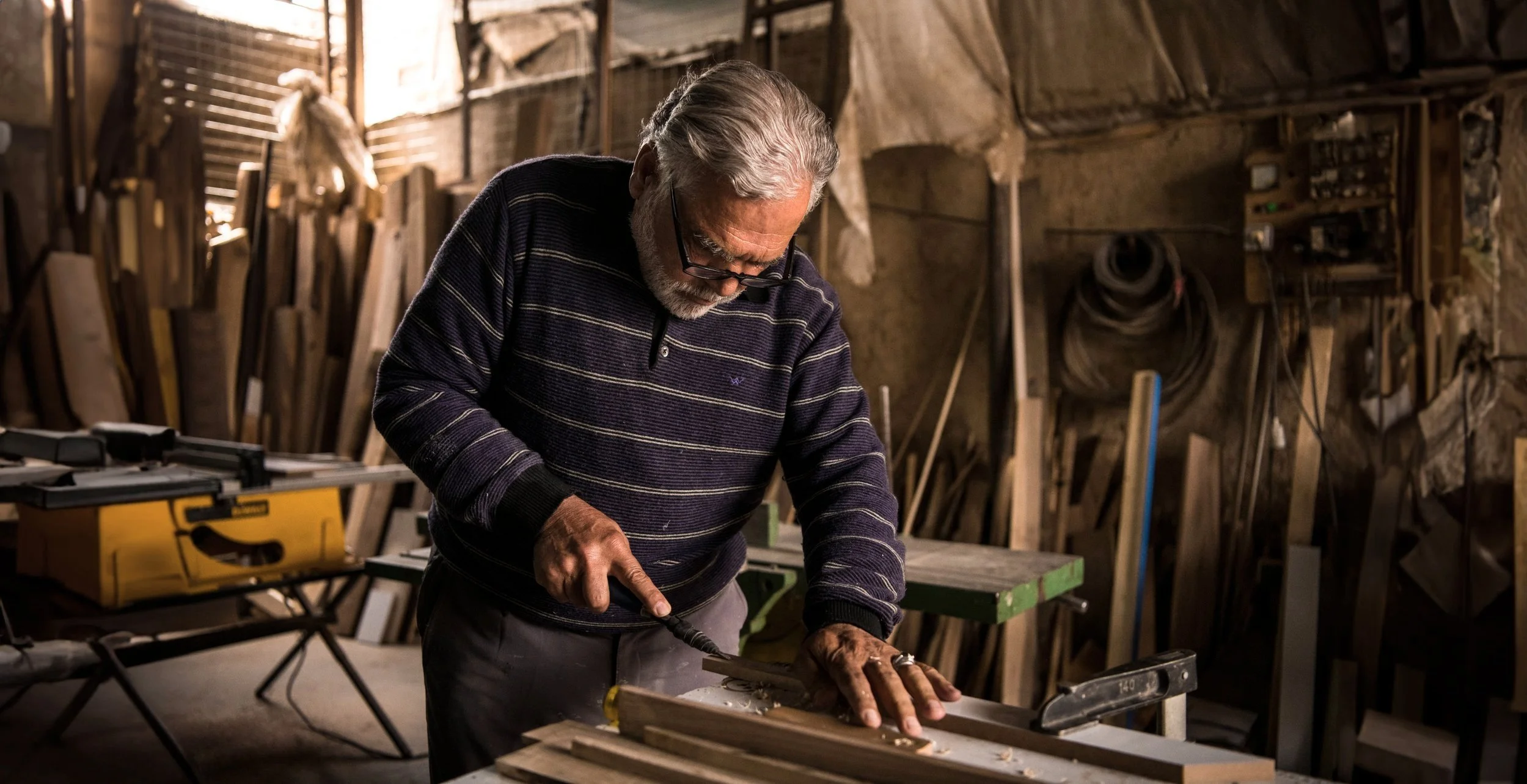 An elderly man with gray hair and beard working on a woodworking project in a workshop, using a hand saw on a piece of wood.