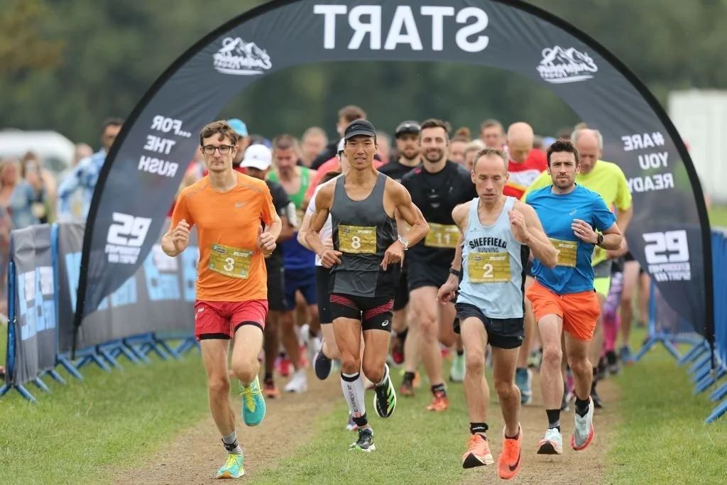 A group of runners at the start of a race, running beneath a black archway with the word 'START' on it, during daytime.