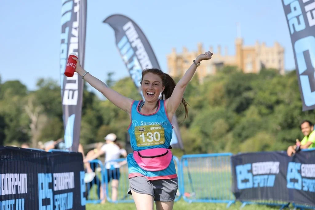Young woman running a marathon, smiling, with her arms raised and holding a red cup, wearing a blue race shirt and grey shorts, at the finish line with flags and other runners in the background on a sunny day.
