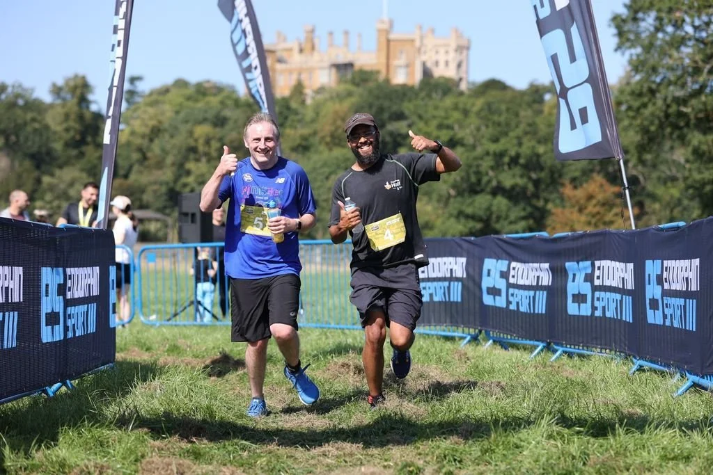 Two men jogging and smiling at a race event, with medals around their necks, holding bottles, giving thumbs up, on a grassy pathway with banners and other runners, in front of a scenic castle and trees.