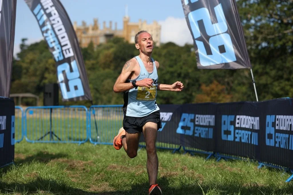 A male runner wearing a light blue tank top and black shorts is crossing the finish line in a race on a grassy field. He has a watch on his left wrist and race bib number 2. There are blue barriers and flags with the logo 'European Sport' in the back