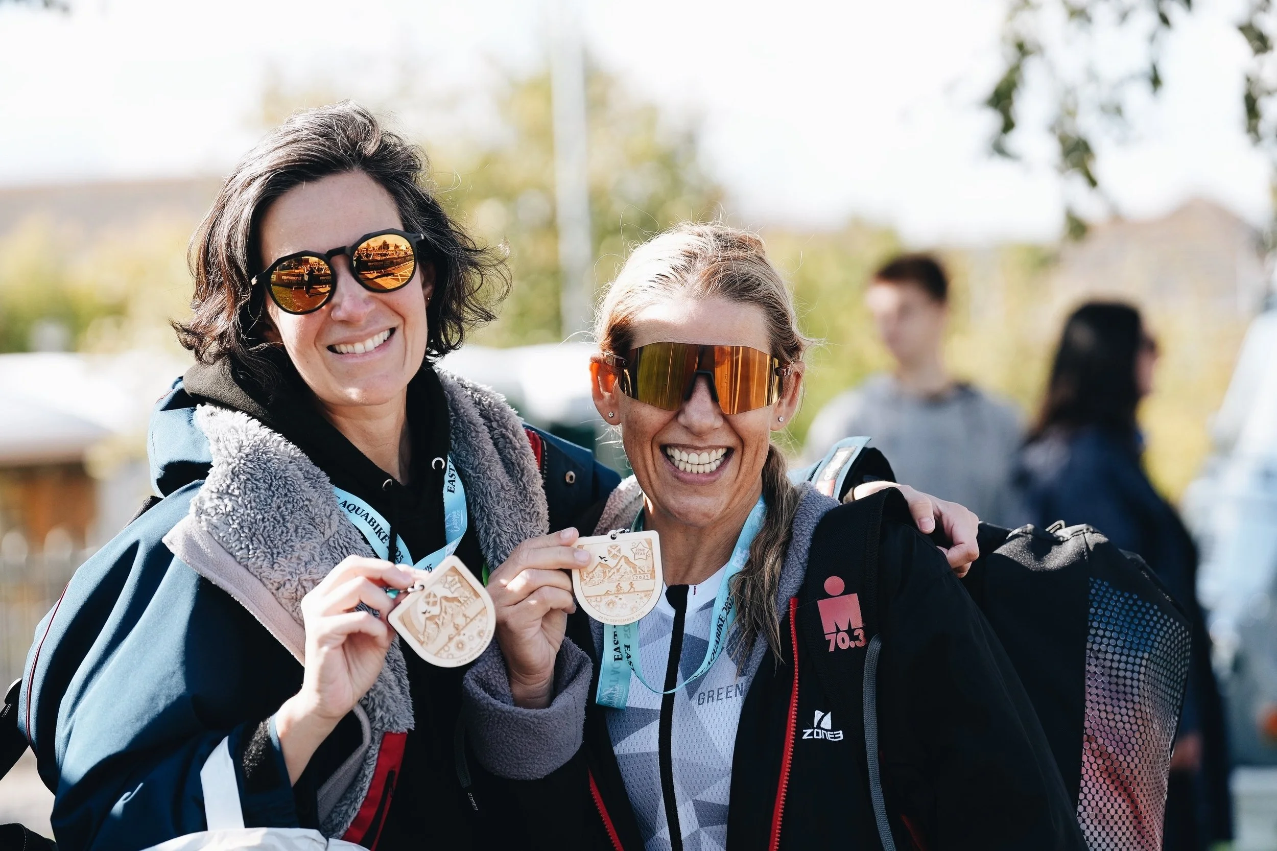 Two women smiling and holding medals outdoors during daytime, wearing sunglasses and outdoor gear, with blurred people and trees in background.