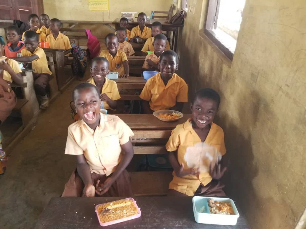 Children wearing school uniforms, sitting in a classroom, smiling and holding lunch containers.