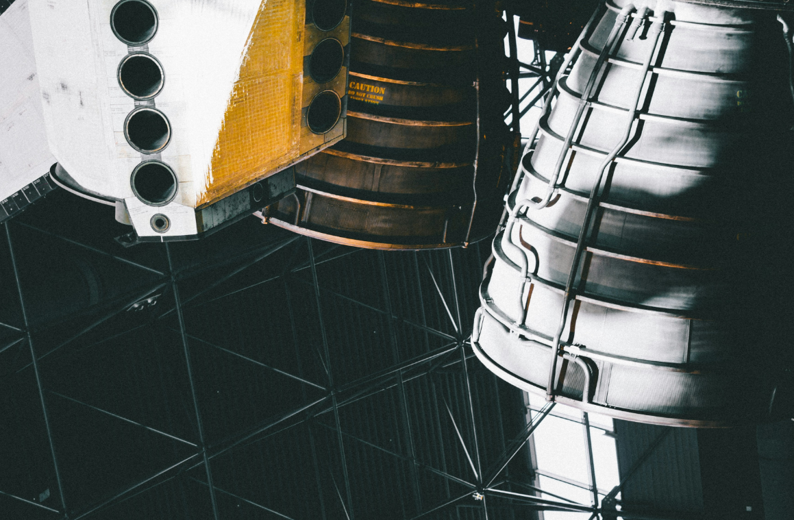 Close-up of spacecraft engines with intricate metallic and black components, situated inside a modern building with a structured black ceiling.