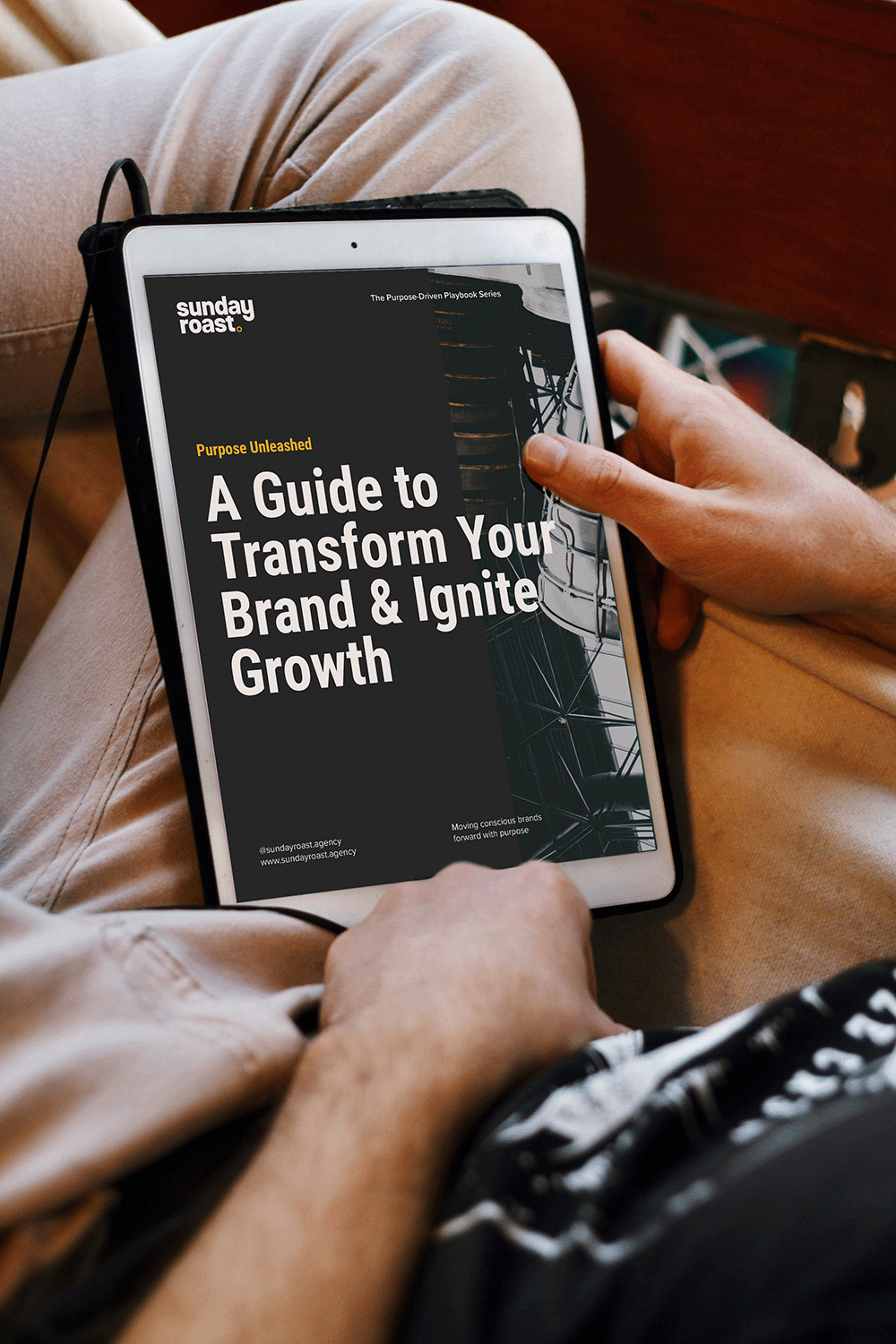 A person holding a tablet displaying a guide titled "A Guide to Transform Your Brand & Ignite Growth" from Sunday Roast agency, sitting on a beige sofa with their legs crossed.