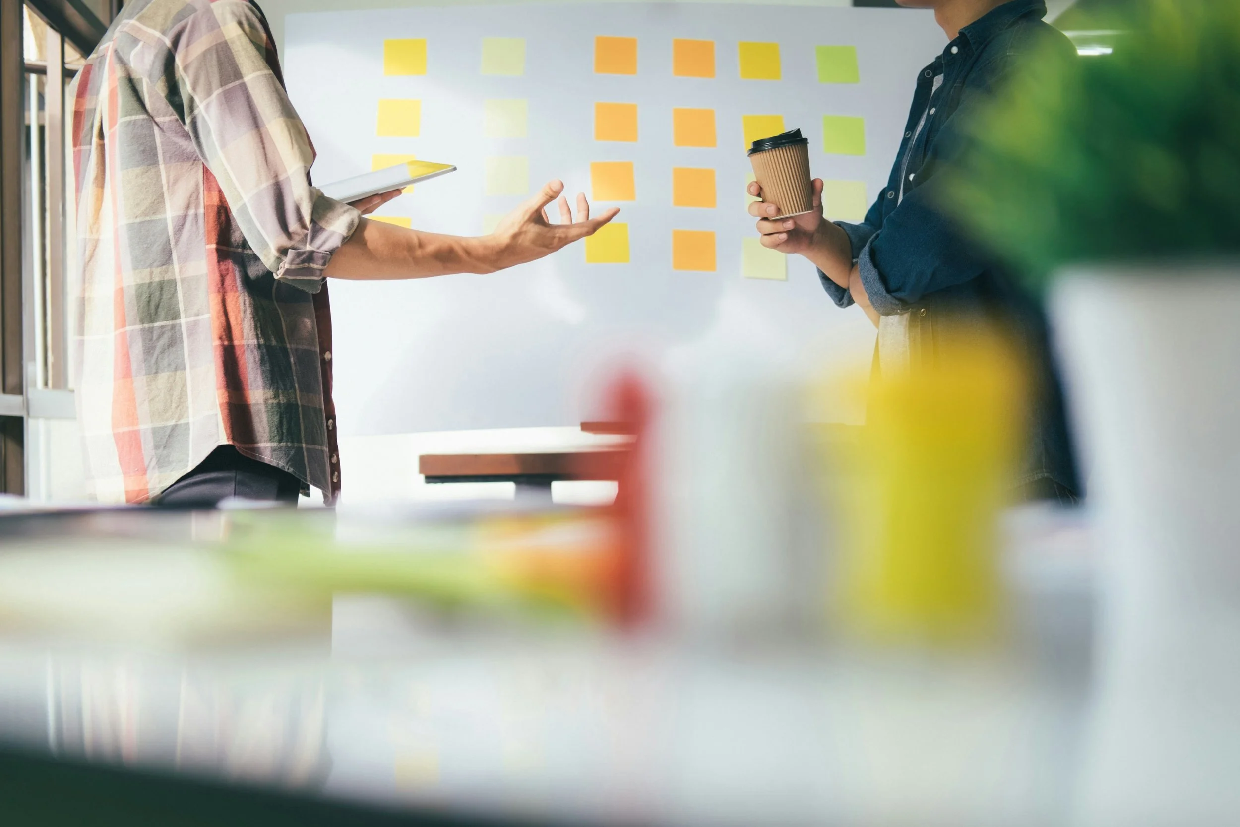 Two people standing in front of a whiteboard covered with colorful sticky notes, one person gesturing with their hand while the other holds a coffee cup, in an office or meeting room.