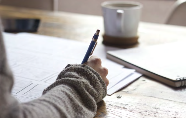 Close-up of a person writing with a pen on papers at a wooden table, with a mug and a closed notebook in the background.