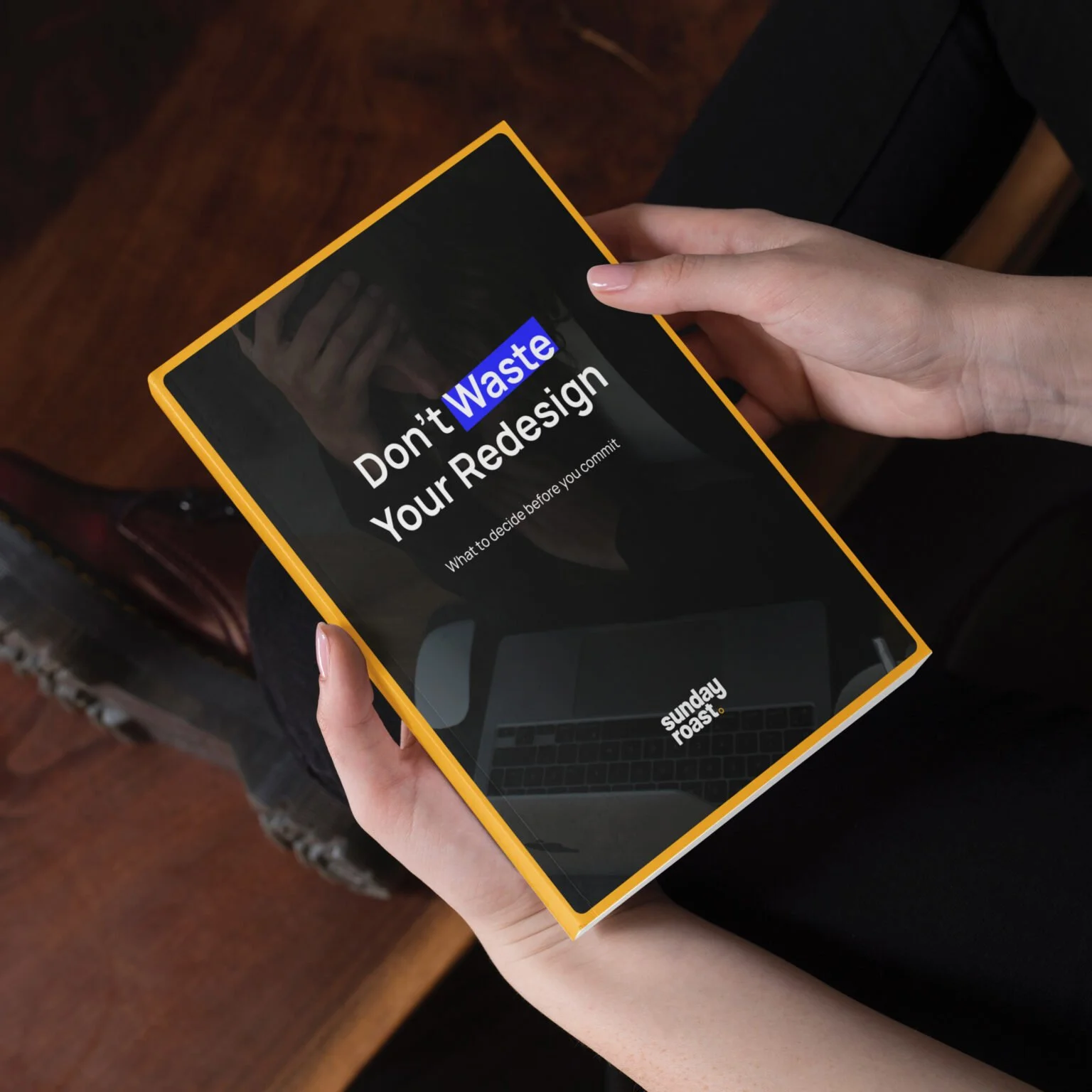 Person holding a black book or magazine with a yellow border that has the title 'Don't Waste Your Redesign' on the cover. The cover also has the text 'What to decide before you commit' and a small logo or text at the bottom that says 'sunday roast'.