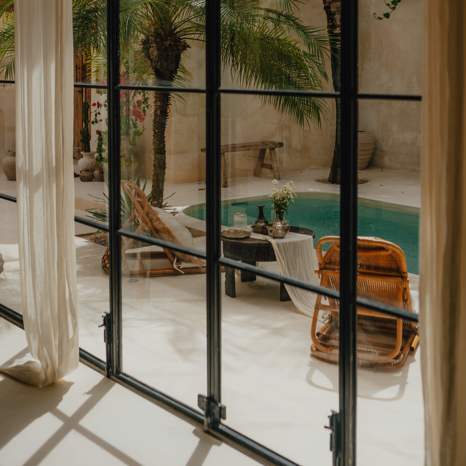 View of a backyard patio area with a small pool, wicker chairs, potted plants, a wooden bench, and large green palm trees, seen through a glass window with black framing.