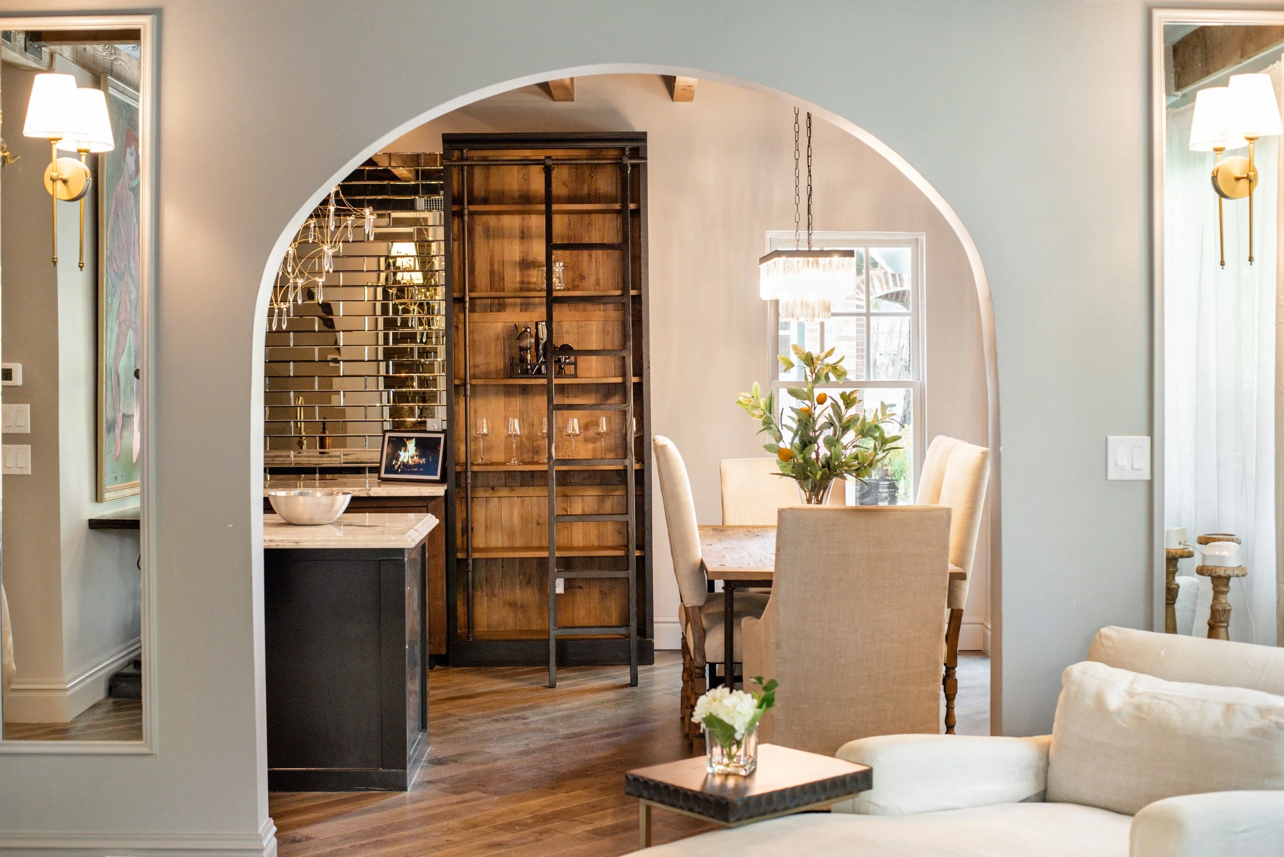 Dining area with beige chairs, a wooden table with a small plant, and a light fixture hanging over the table, visible through an arched opening.
