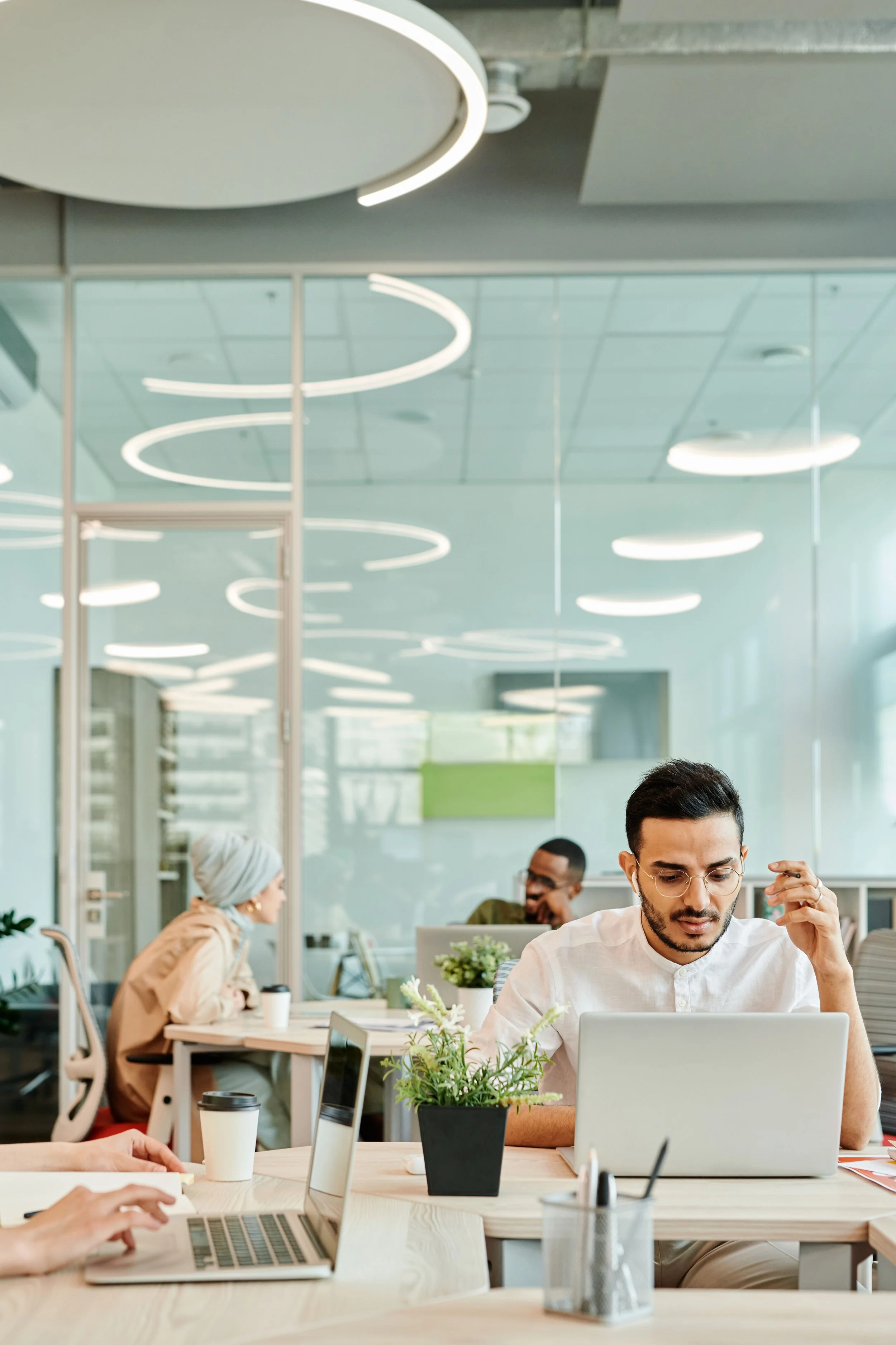A diverse group of office workers working at desks in a modern open-plan office with glass walls and ceiling lights.