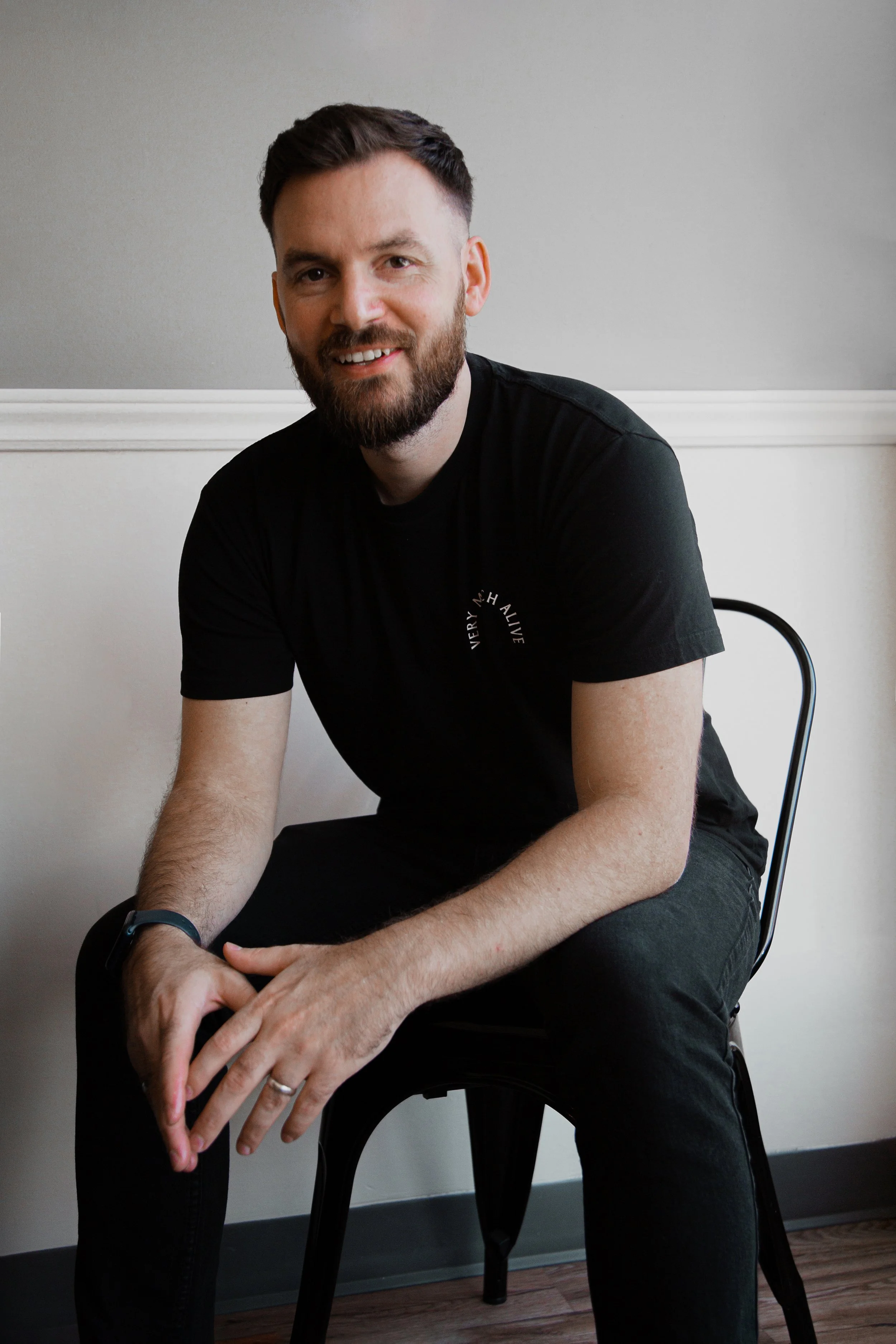 A bearded man wearing a black T-shirt, sitting on a black chair against a plain gray wall, smiling at the camera.