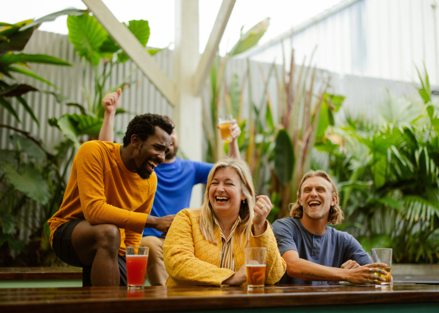 Group of friends laughing and having drinks outdoors surrounded by lush green plants.