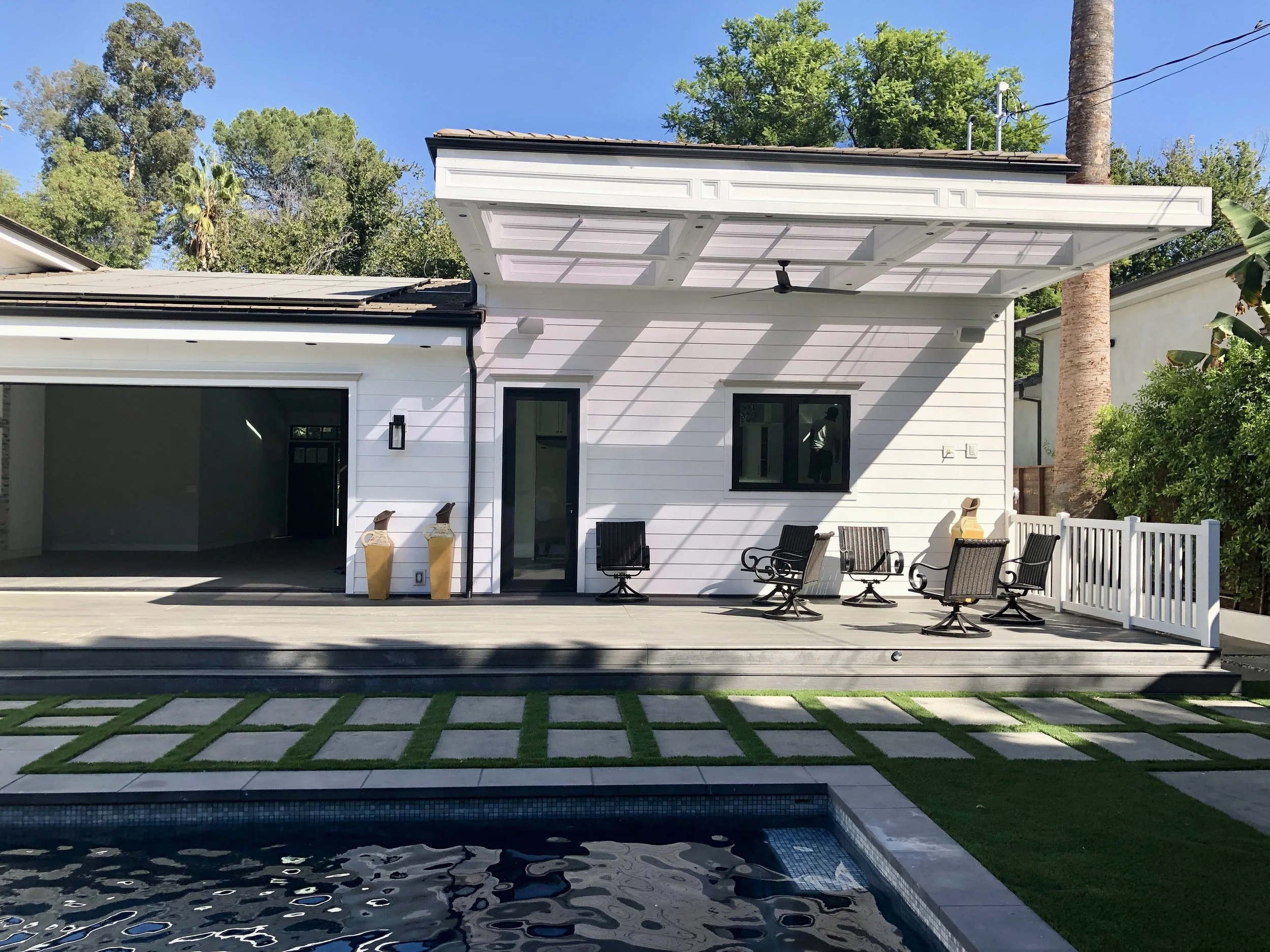 Backyard with a covered patio, outdoor chairs, a pool in the foreground, and a white house with a sliding door and window, surrounded by green trees and clear sky.