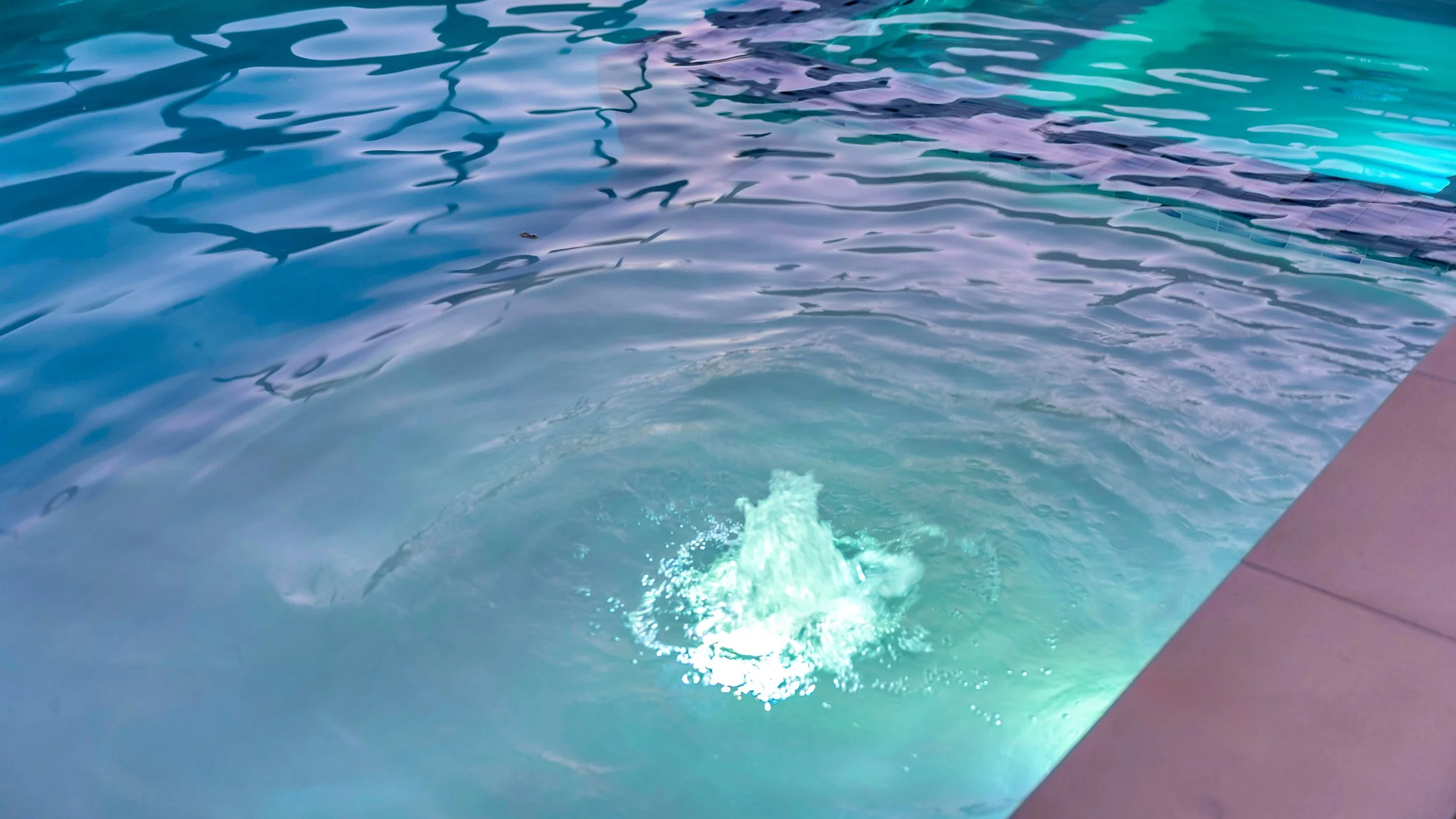 An underwater pool light illuminating the water with a bright glow, near the edge of a swimming pool with blue water and a purple border.