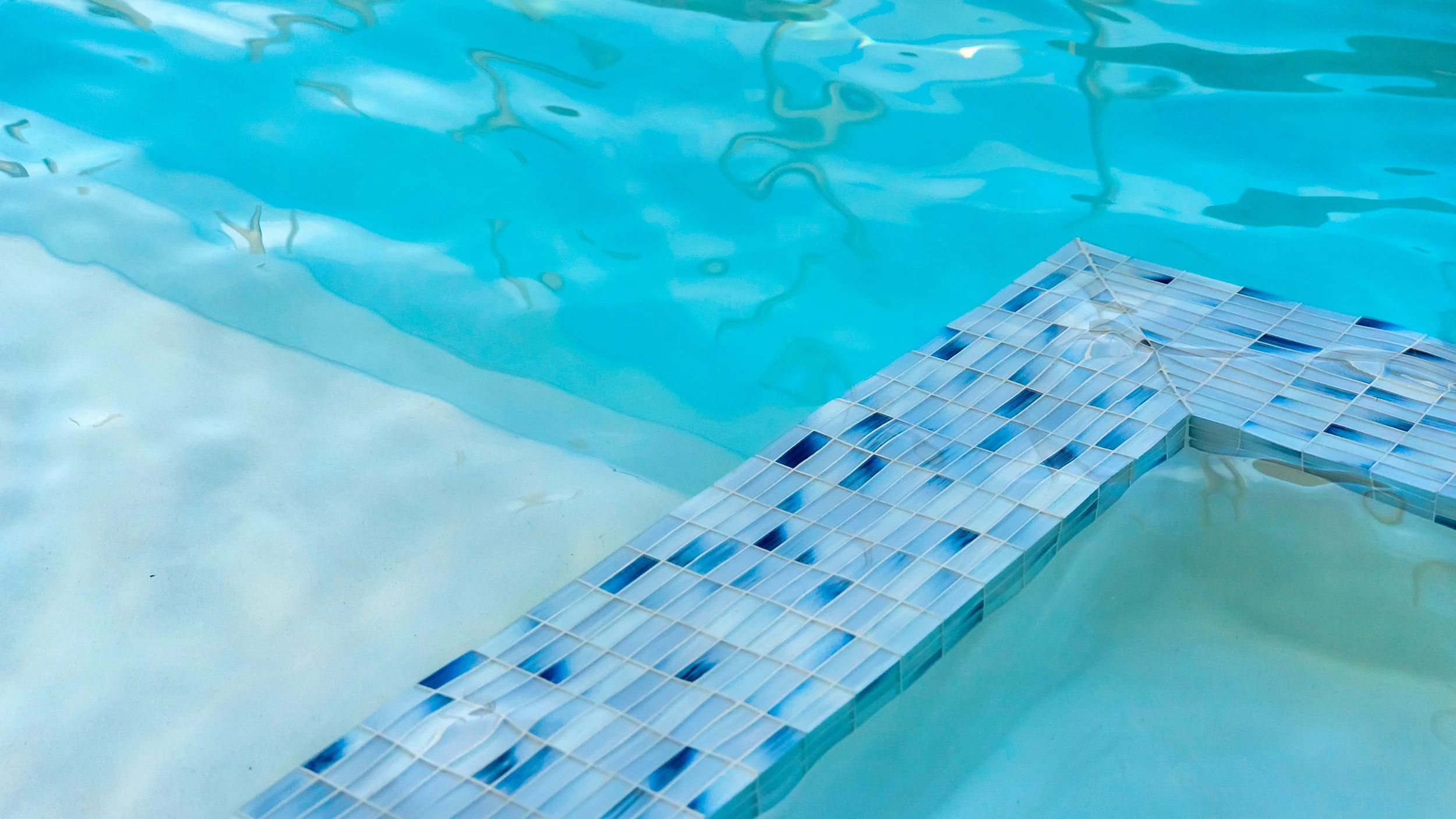 Close-up of a swimming pool with a mosaic tile ledge in the water, showing blue tiles and the rippling surface of the pool.