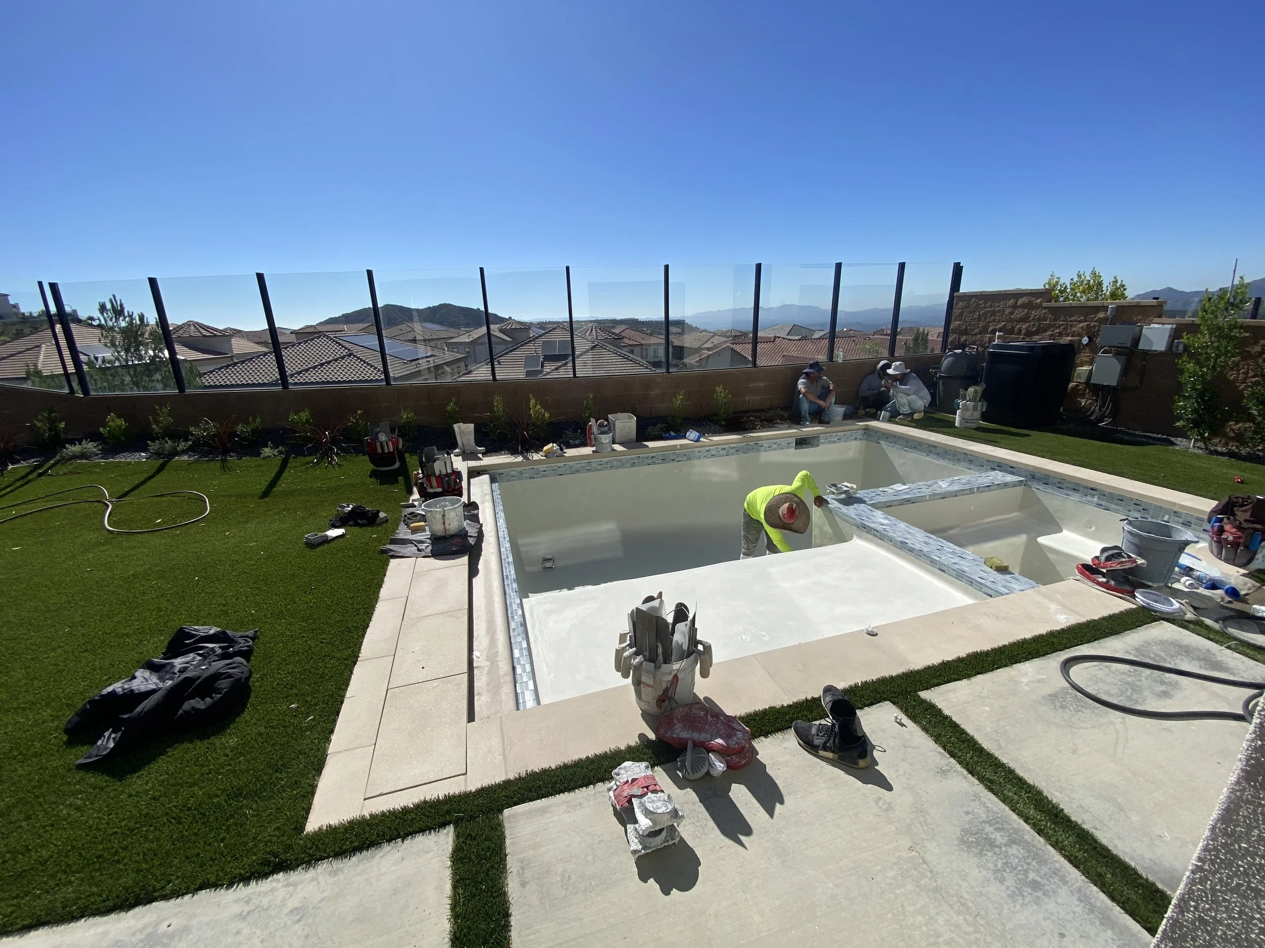 Workers installing a pool in a backyard with green artificial grass and a glass fence, across rooftops, mountains, and a clear blue sky in the background.