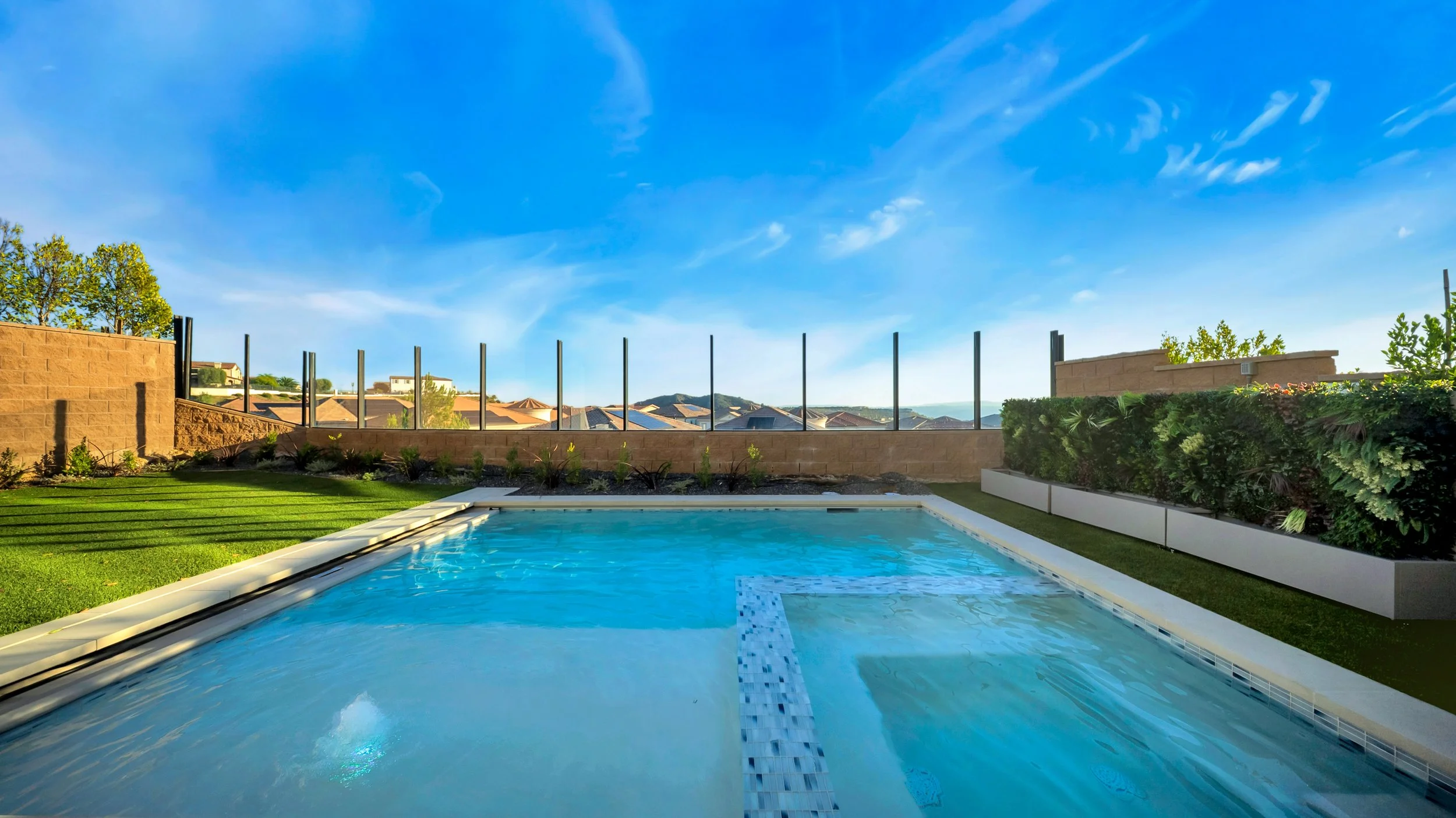 View of a backyard swimming pool with a clear blue sky above, surrounded by a green lawn, a brick wall, and glass fence panels, with neighboring houses and trees in the distance.