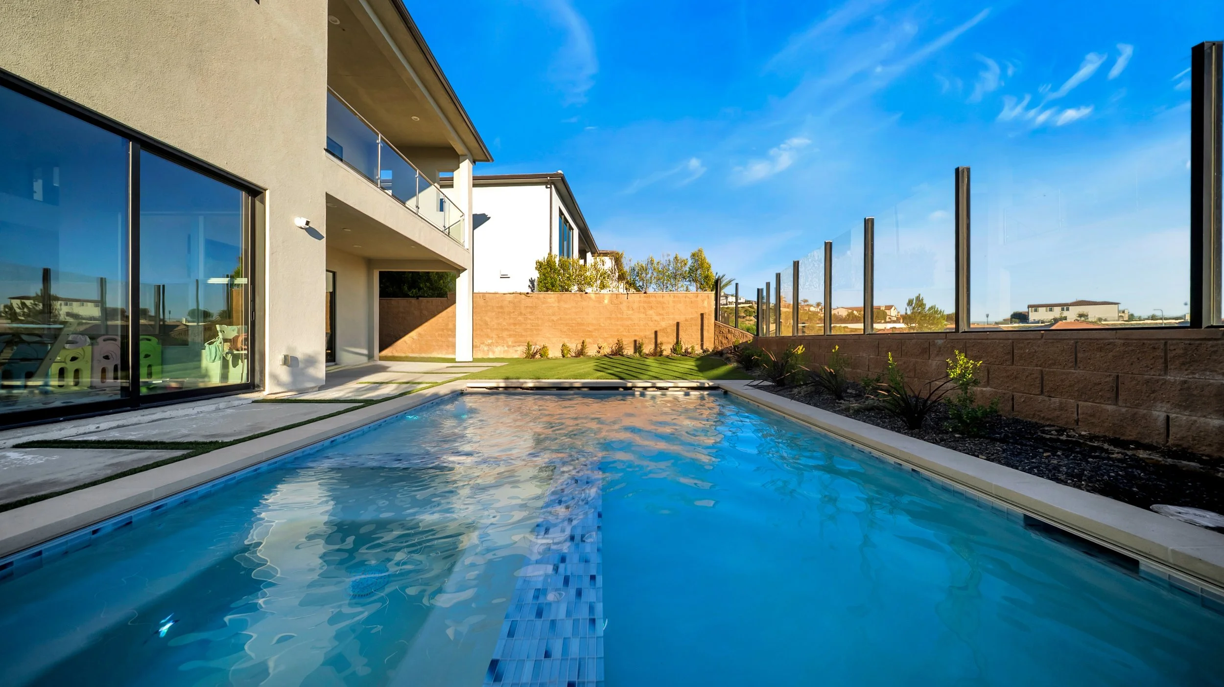 Modern backyard with a swimming pool next to a house, glass sliding doors, a small lawn, plants, and a glass fence under a clear blue sky.
