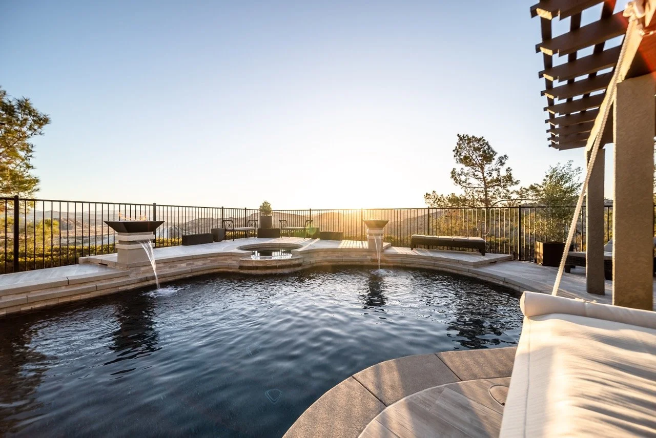 A swimming pool with waterfall features, surrounded by a metal railing, with a scenic sunset view of trees and hills in the background, and outdoor seating areas nearby.