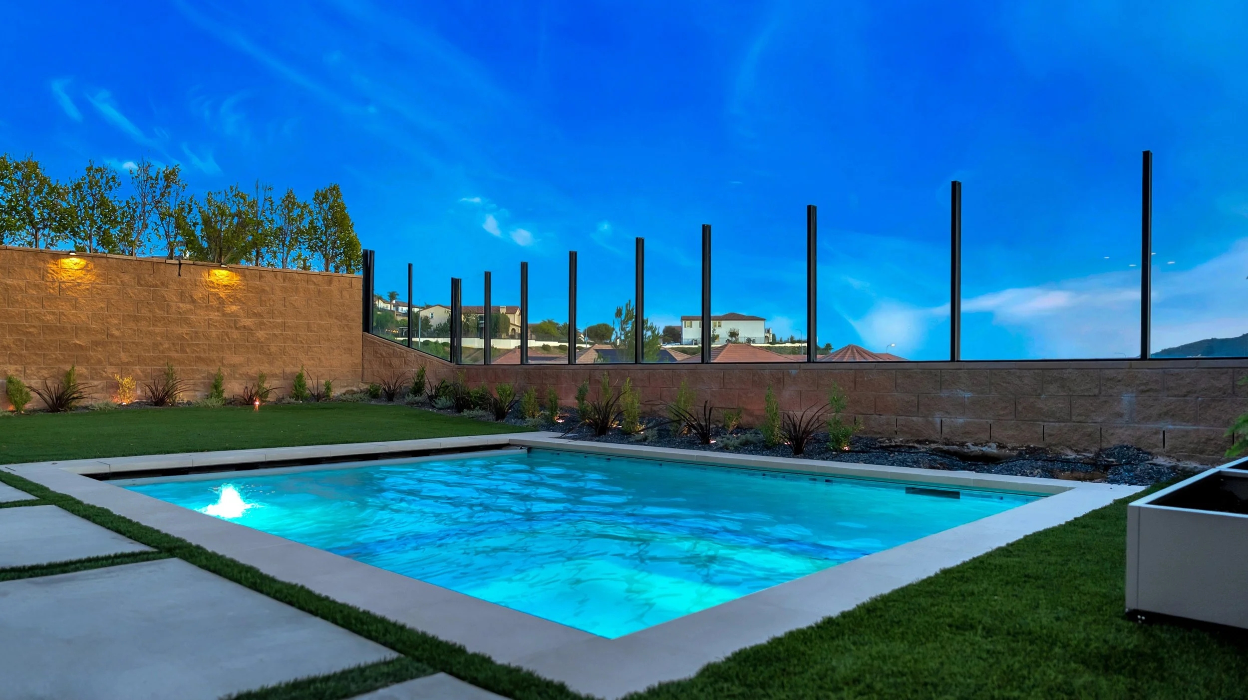 Residential backyard with a small swimming pool illuminated, surrounded by green lawn and decorative plants, under a clear blue evening sky.