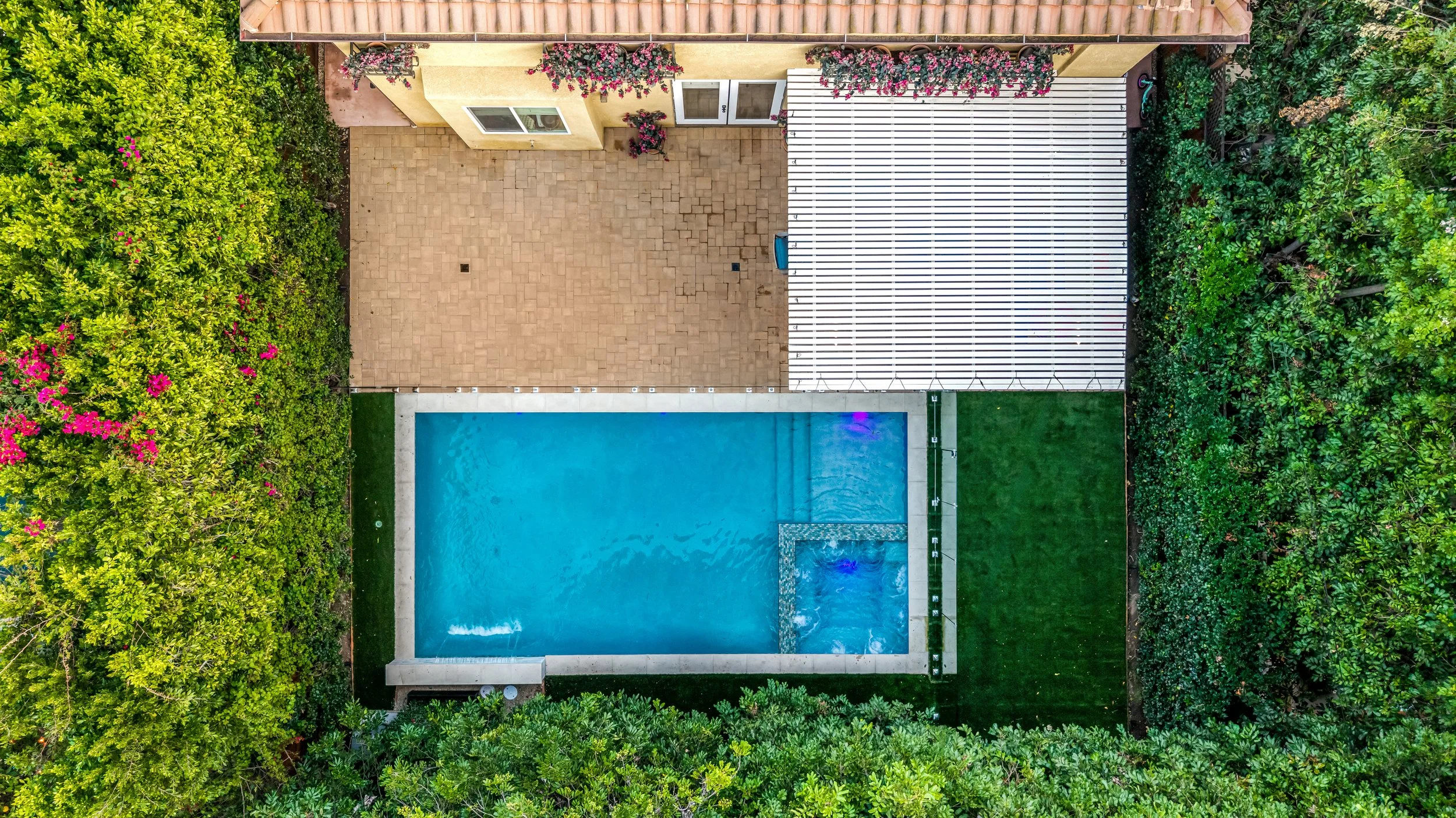 Aerial view of a backyard with a swimming pool, surrounded by lush green trees and plants, featuring a paved patio area, a white pergola, and a grassy area.
