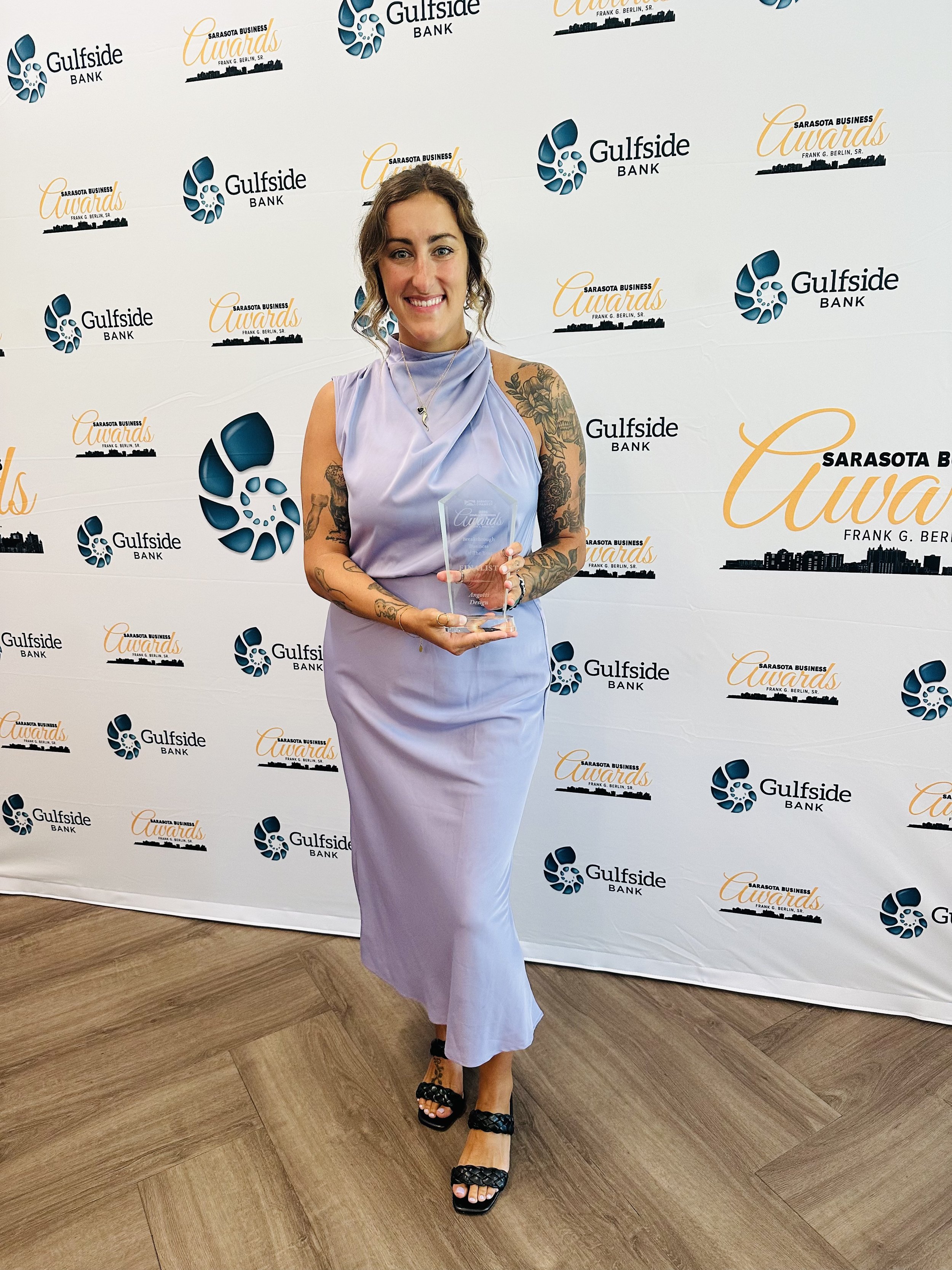 Frankie Angotti in a lavender dress holding an award at an event with a backdrop featuring the logos for Gulfside Bank and the Sarasota Business Awards.