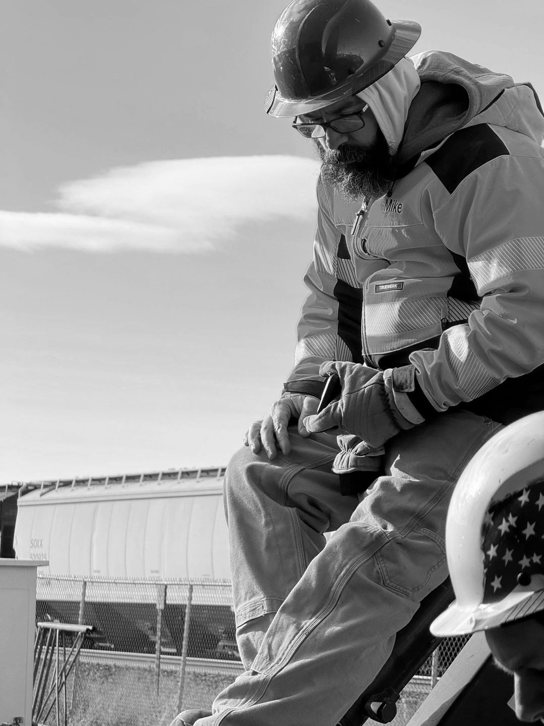 A man with a beard wearing a safety helmet, glasses, and a bright yellow high-visibility jacket sits outdoors on a white chair, with a large shipping container and a clear blue sky in the background.