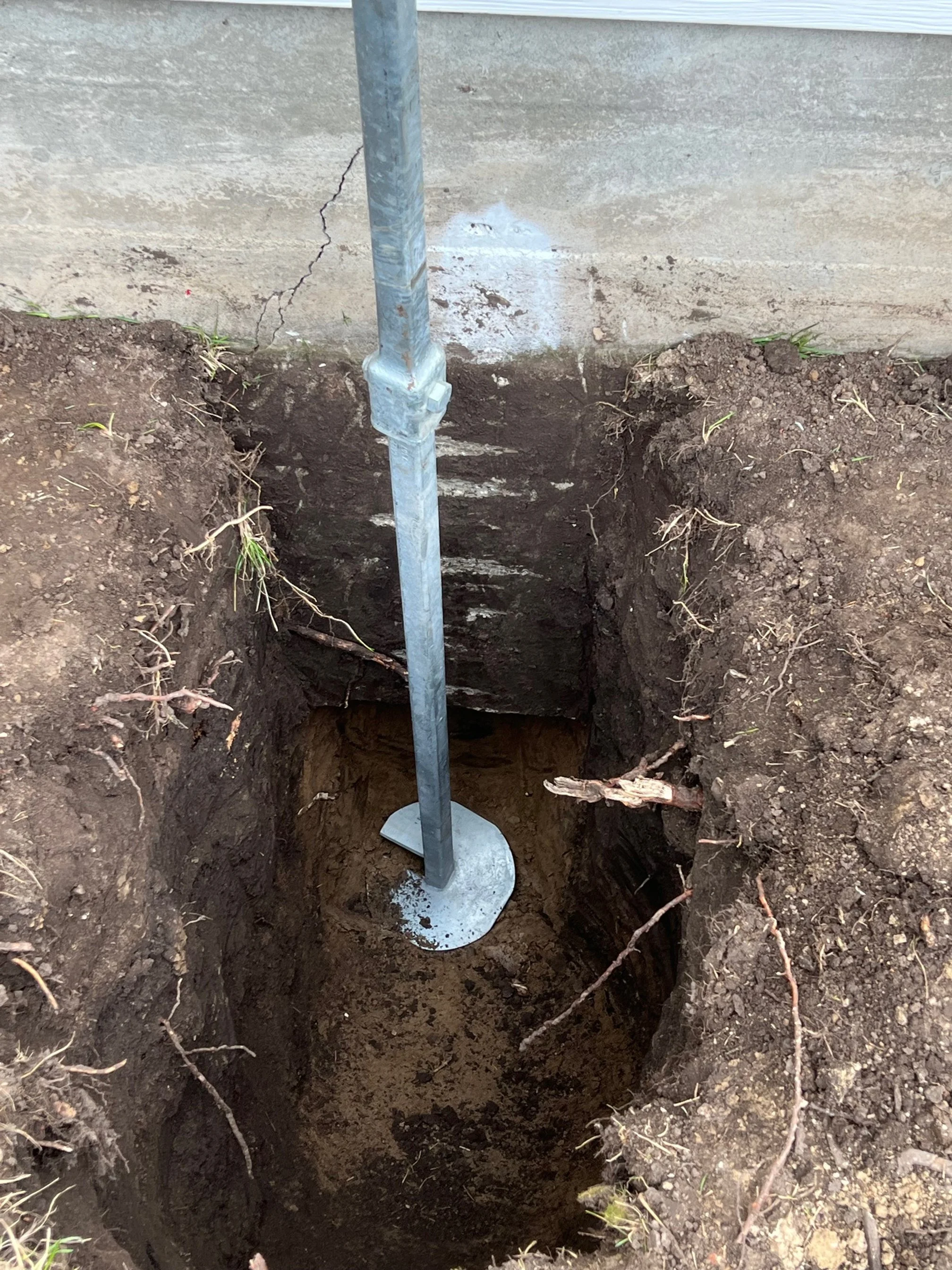 A metal post stands upright in a deep hole dug into the soil adjacent to a concrete wall.