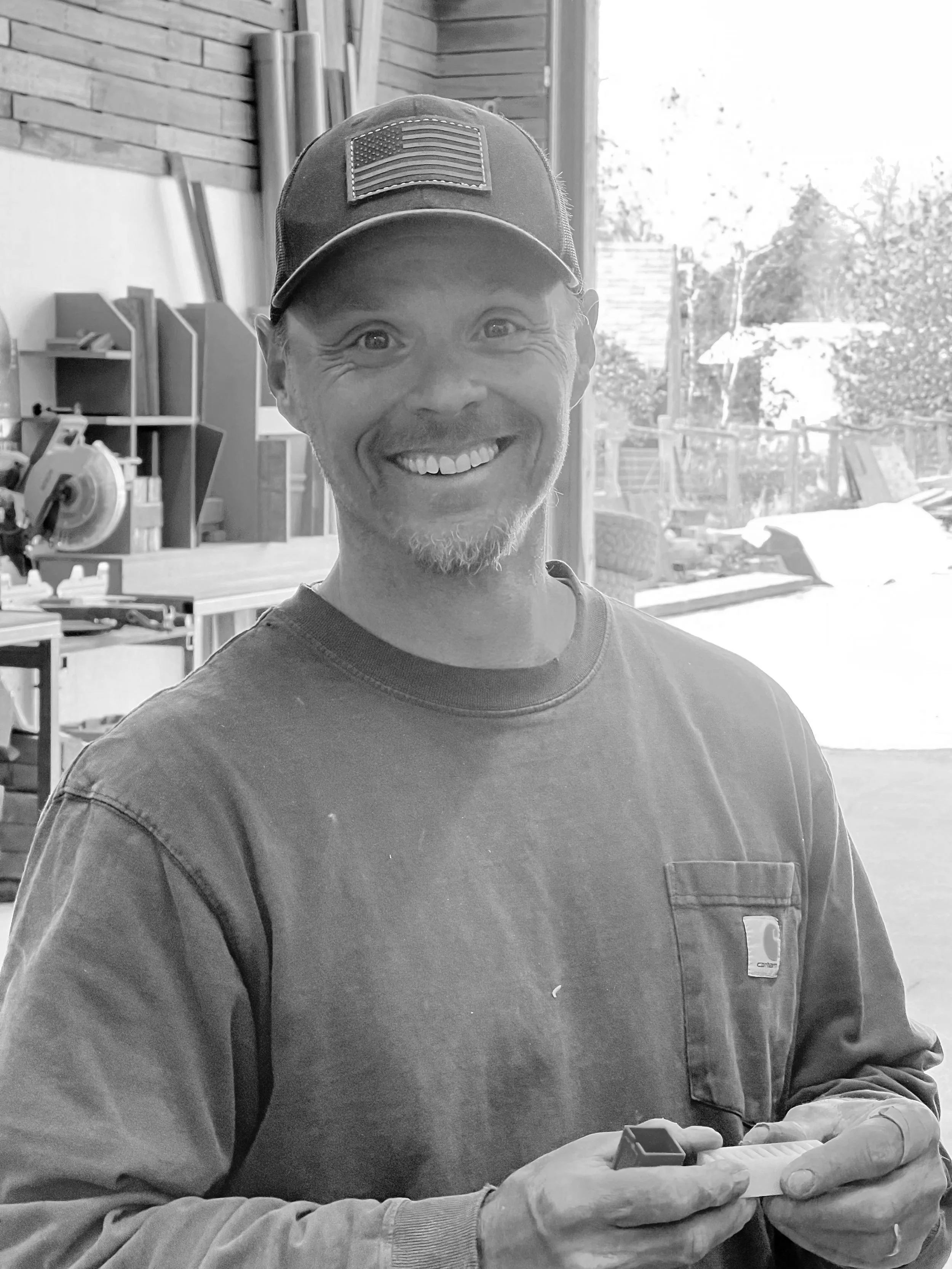 A smiling man wearing a black cap with an American flag patch and a dark gray Carhartt long sleeve shirt, holding a small red object in his hands, inside a woodworking shop with tools and wooden materials in the background.