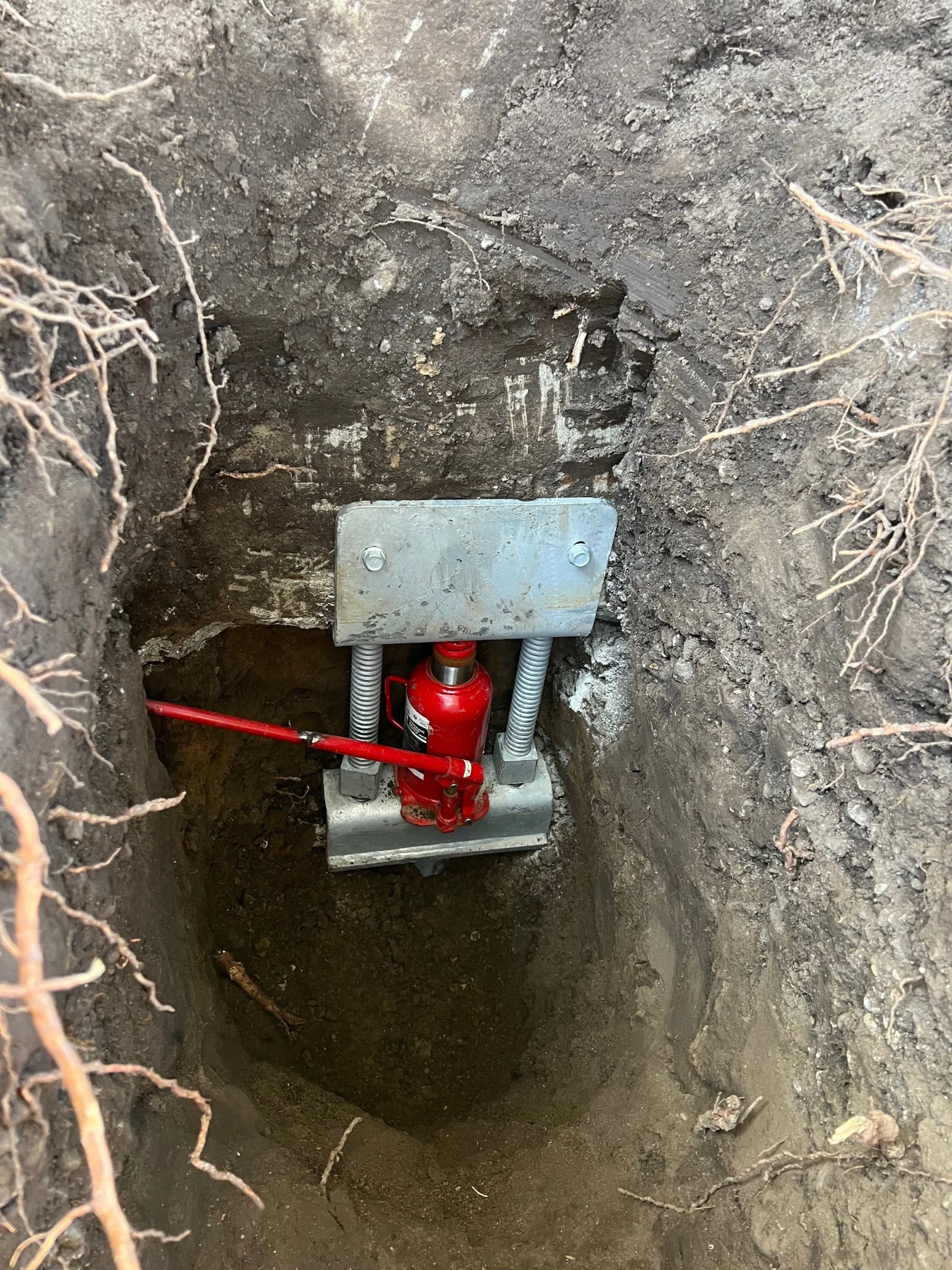 A metal hydraulic or pneumatic jack installed at the bottom of a deep hole with exposed soil walls and some small roots.