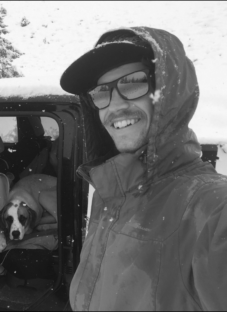 A man in brown waterproof jacket and black sunglasses taking a selfie in a snowy outdoor setting, with a dog lying on the seat inside a vehicle behind him.