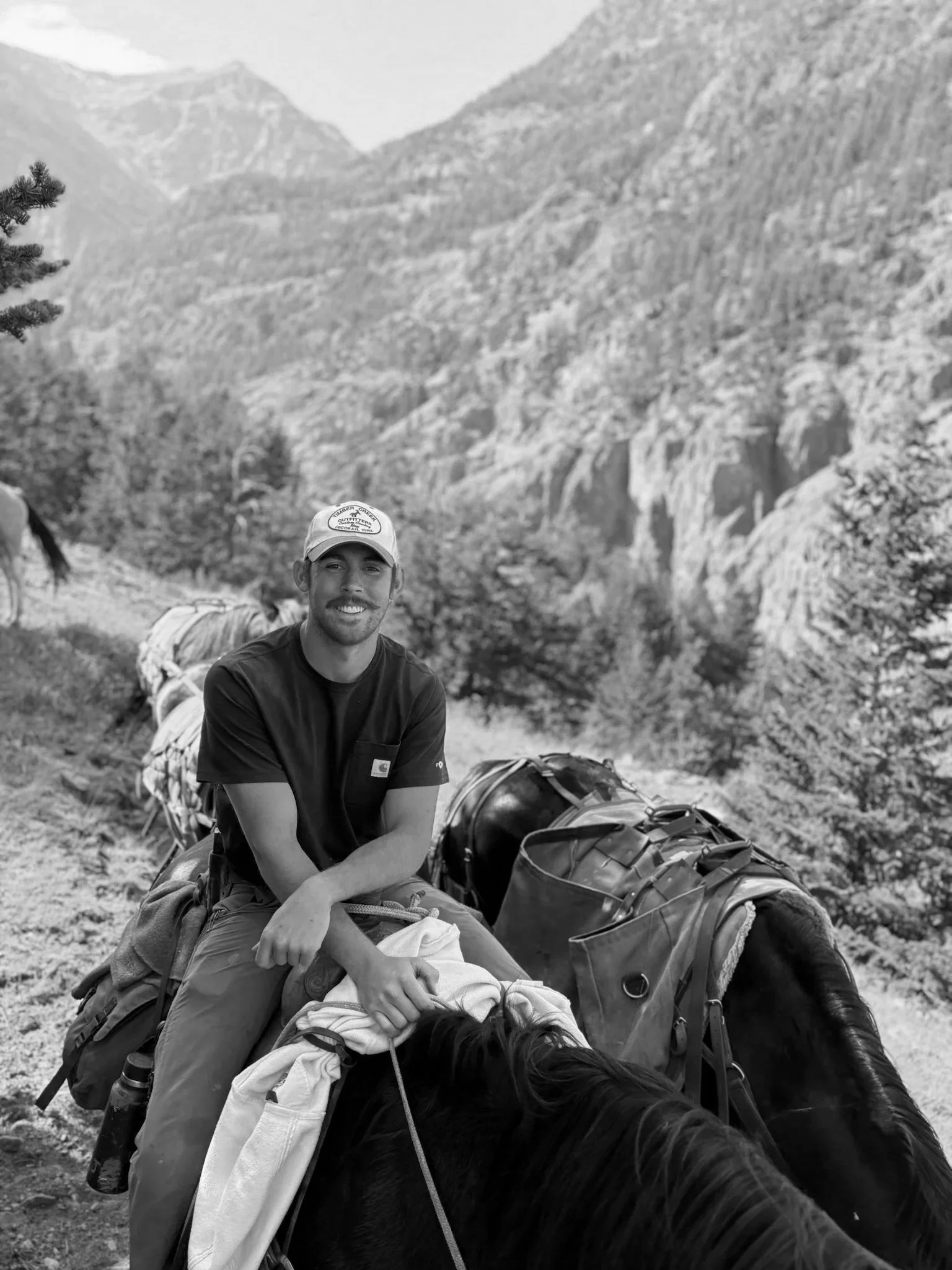A man sitting on the ground in a mountainous area with trees, wearing a cap and black t-shirt, smiling at the camera. There are camping gear and a horse lying on the ground in front of him, with a mountainous landscape in the background.