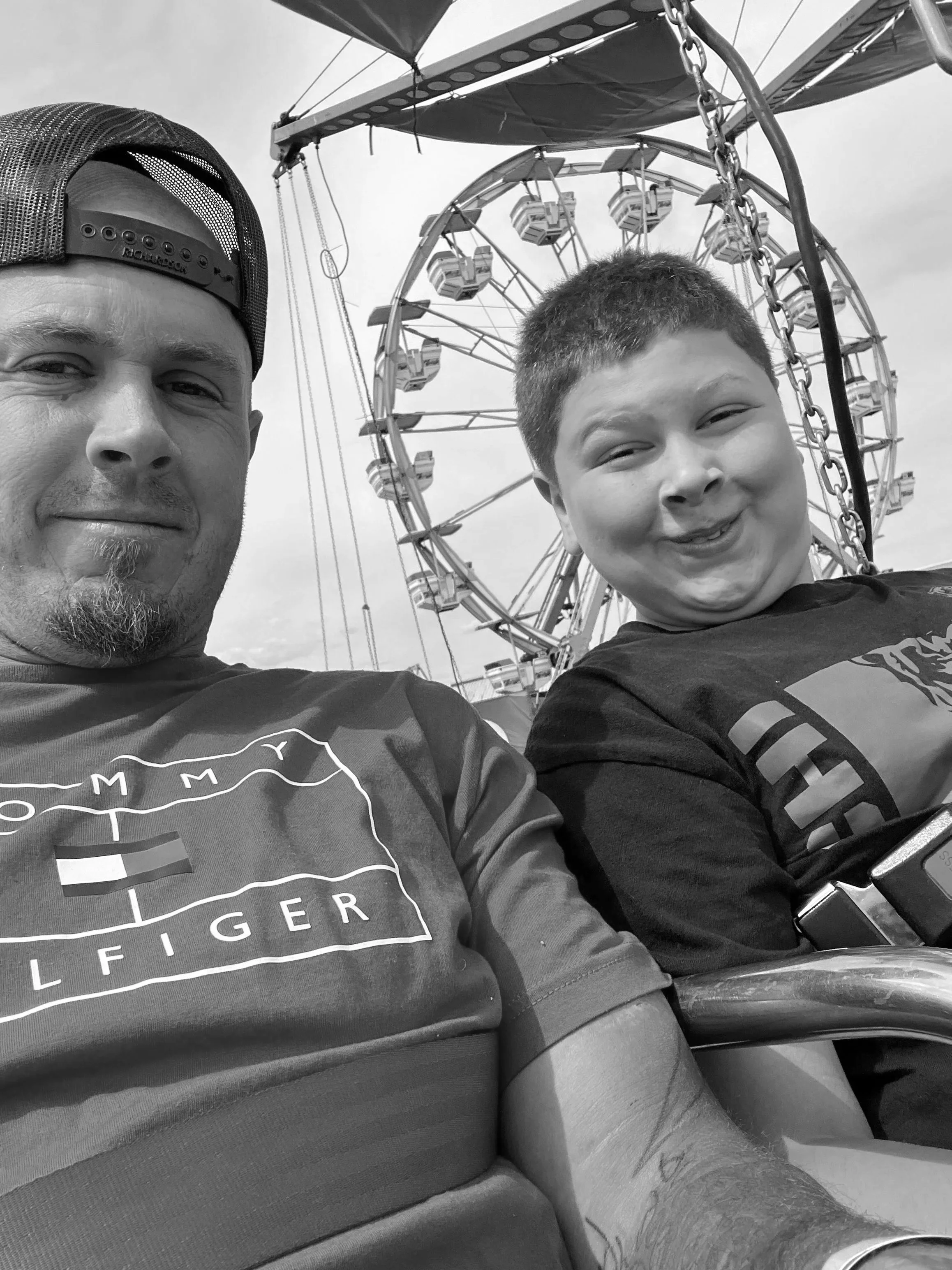 A man and boy taking a selfie at a fairground with a Ferris wheel in the background.