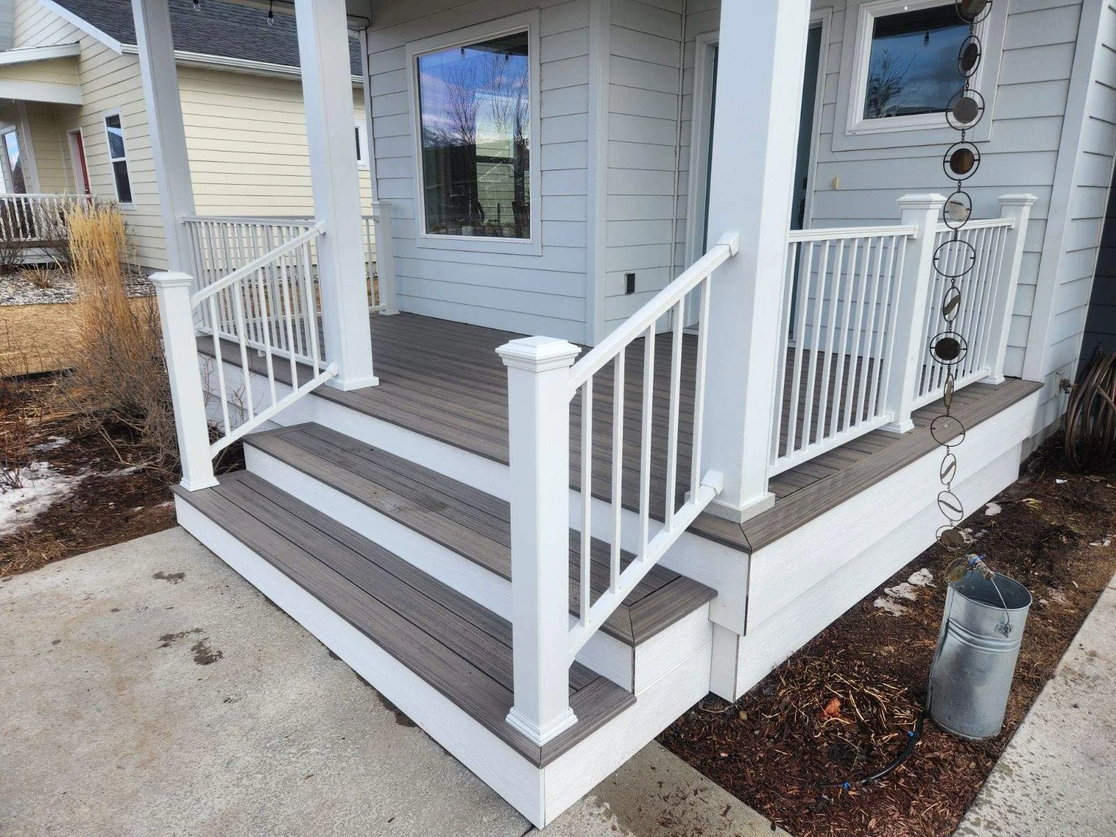 A small wooden porch with white railings and stairs leading down to a concrete walkway, attached to a house with light blue siding and a large window.