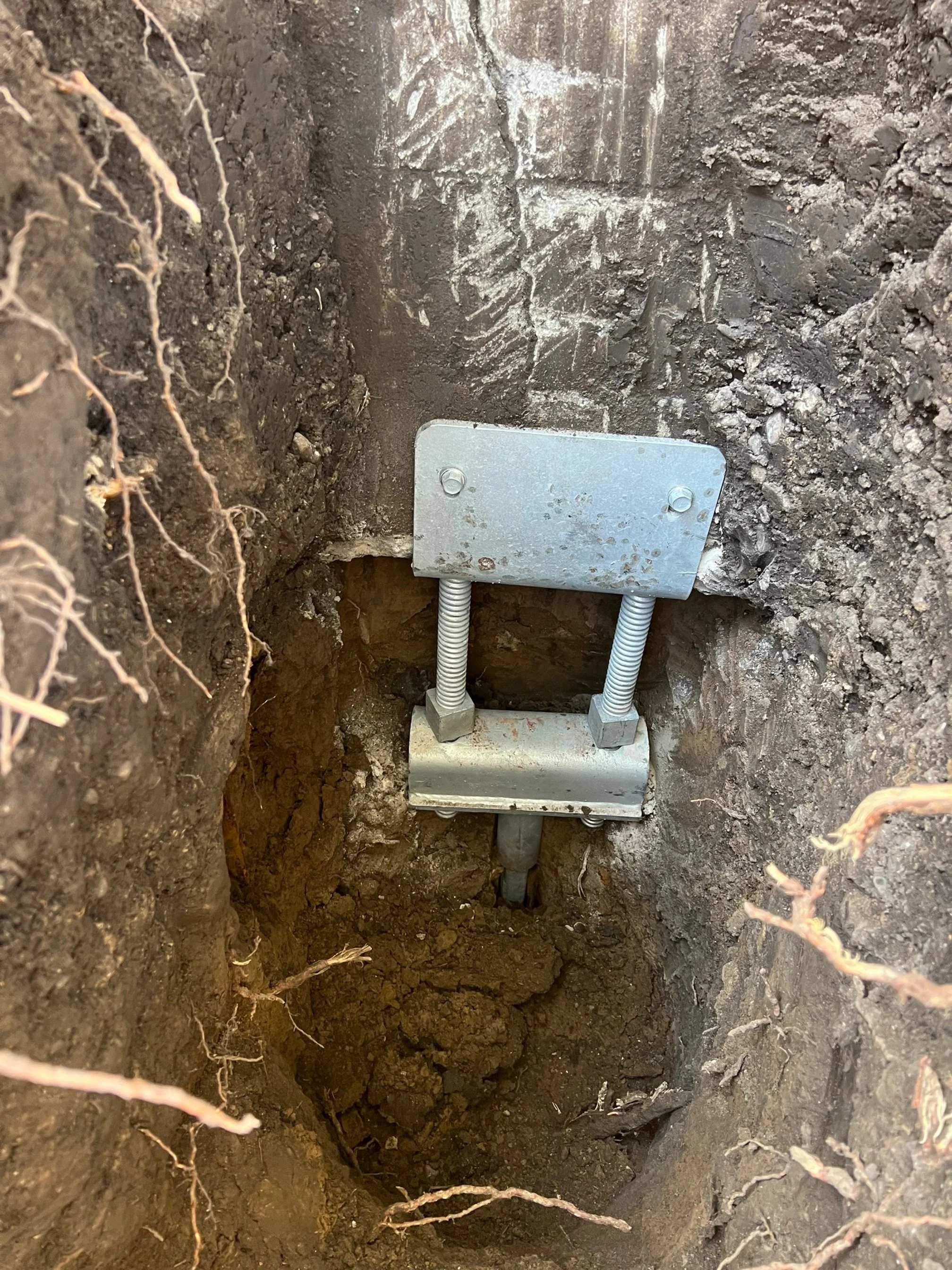 An exposed underground utility junction box with bolts and wiring, embedded in a trench with soil walls.