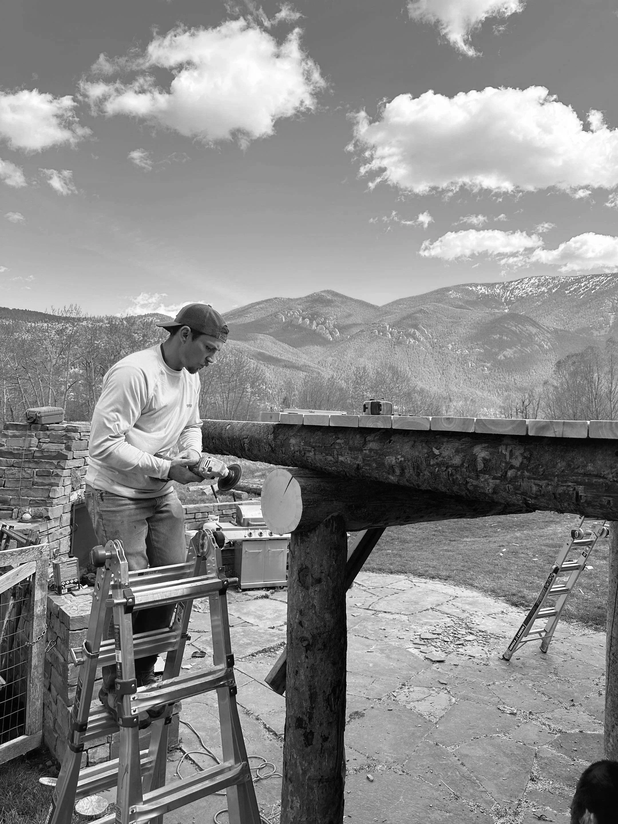 A man wearing a white long sleeve shirt and a backwards gray cap is working with power tools on a large wooden log outdoors. There are ladders, tools, and equipment around him, with a mountain landscape and a blue sky with clouds in the background.