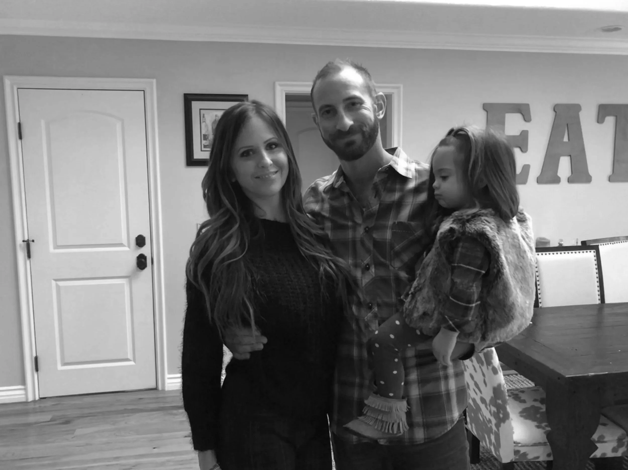 A family of three posing indoors, with a woman, man, and young girl. The woman has long brown hair and is wearing a black sweater. The man has short hair and a beard, wearing a plaid shirt. The young girl has brown hair and is wearing a fur vest and polka dot leggings, being held by the man.