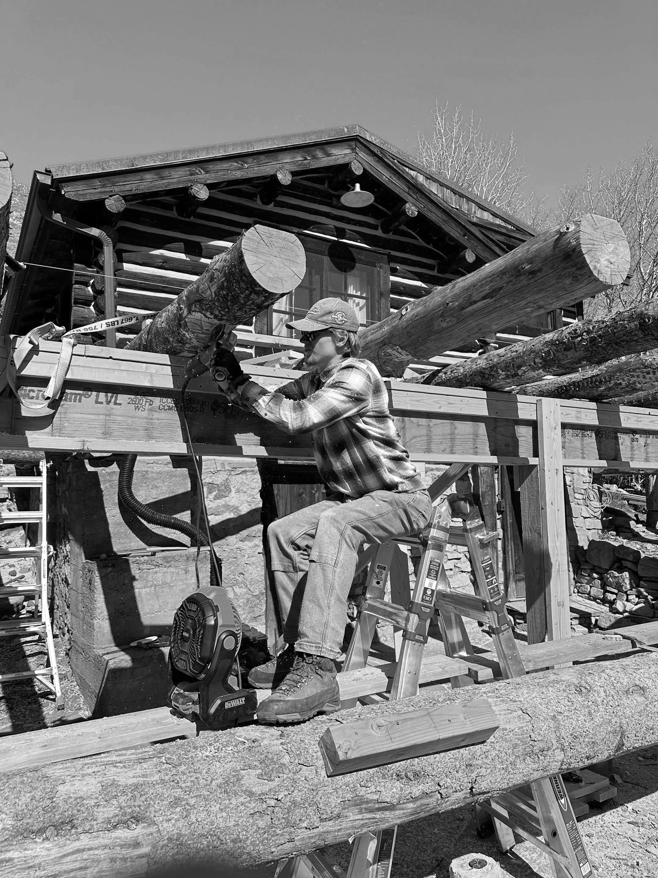 A man working on a construction project outside of a log cabin, using power tools and wearing safety glasses, a baseball cap, and work boots, with ladders and wooden beams around him.