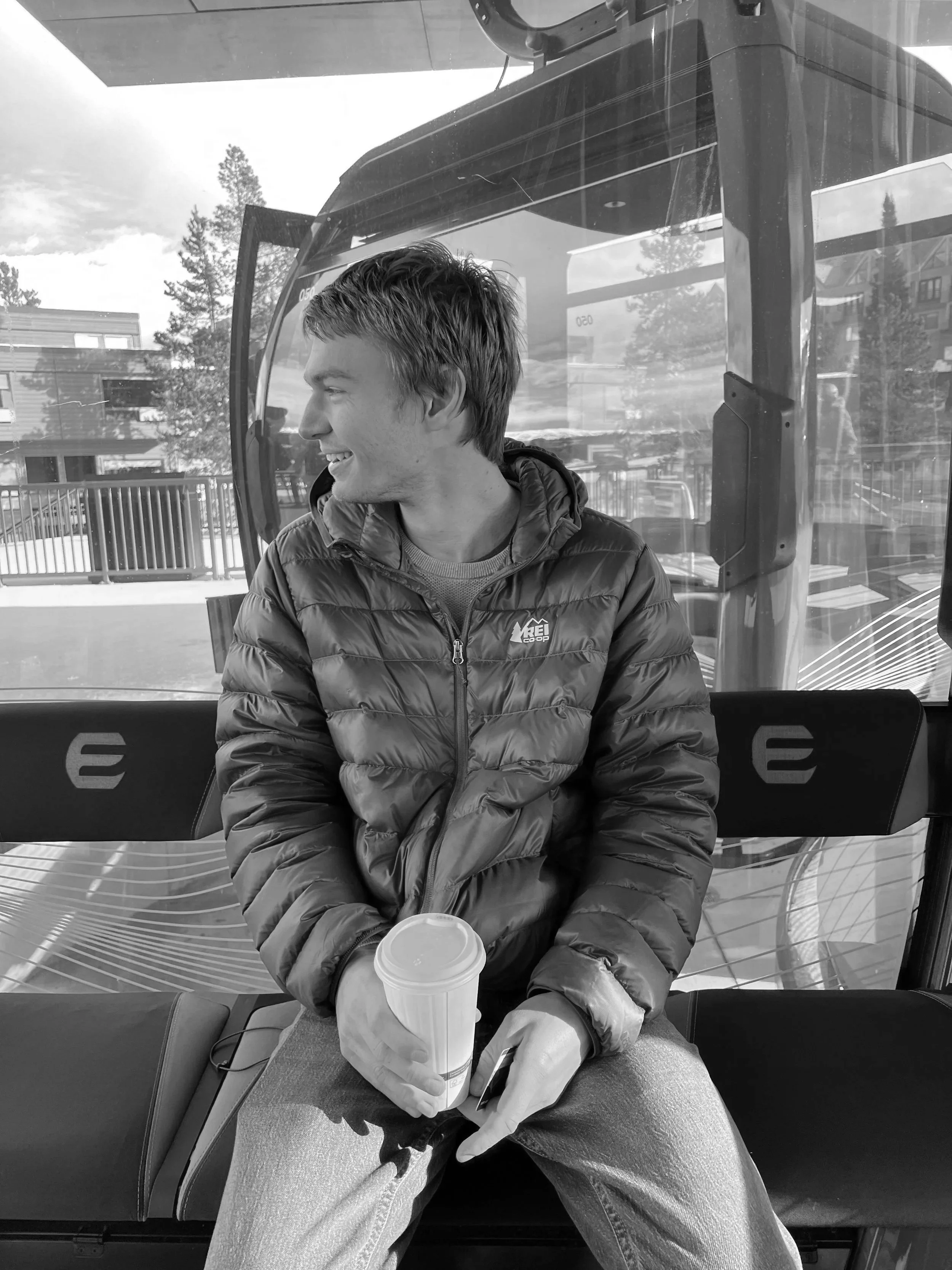 A man wearing a green puffy jacket and blue jeans sitting on a bench at a ski resort, holding a white disposable coffee cup, with snow-covered hills and buildings visible through the window behind him.