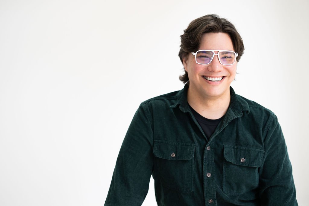 Alex wears a green shirt and white-rimmed glasses. He smiles directly at the camera.