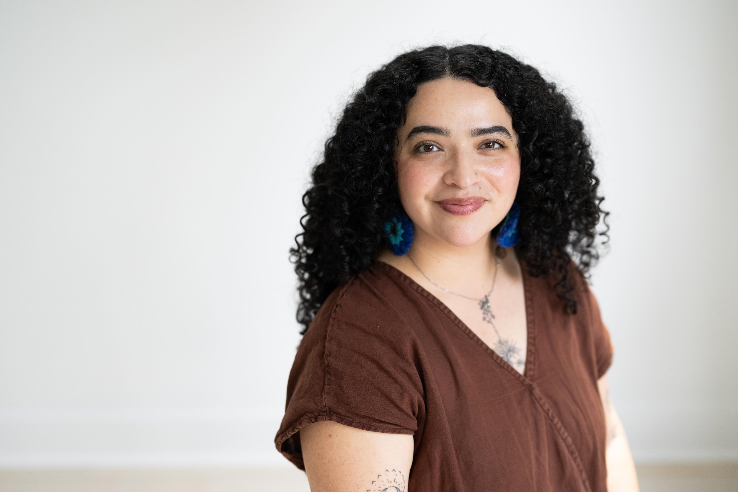 A photo of Karina Monroy, a Chicana woman with curly dark hair who is wearing a brown top and long blue crocheted earrings. She is looking directly into the camera and smiling softly.