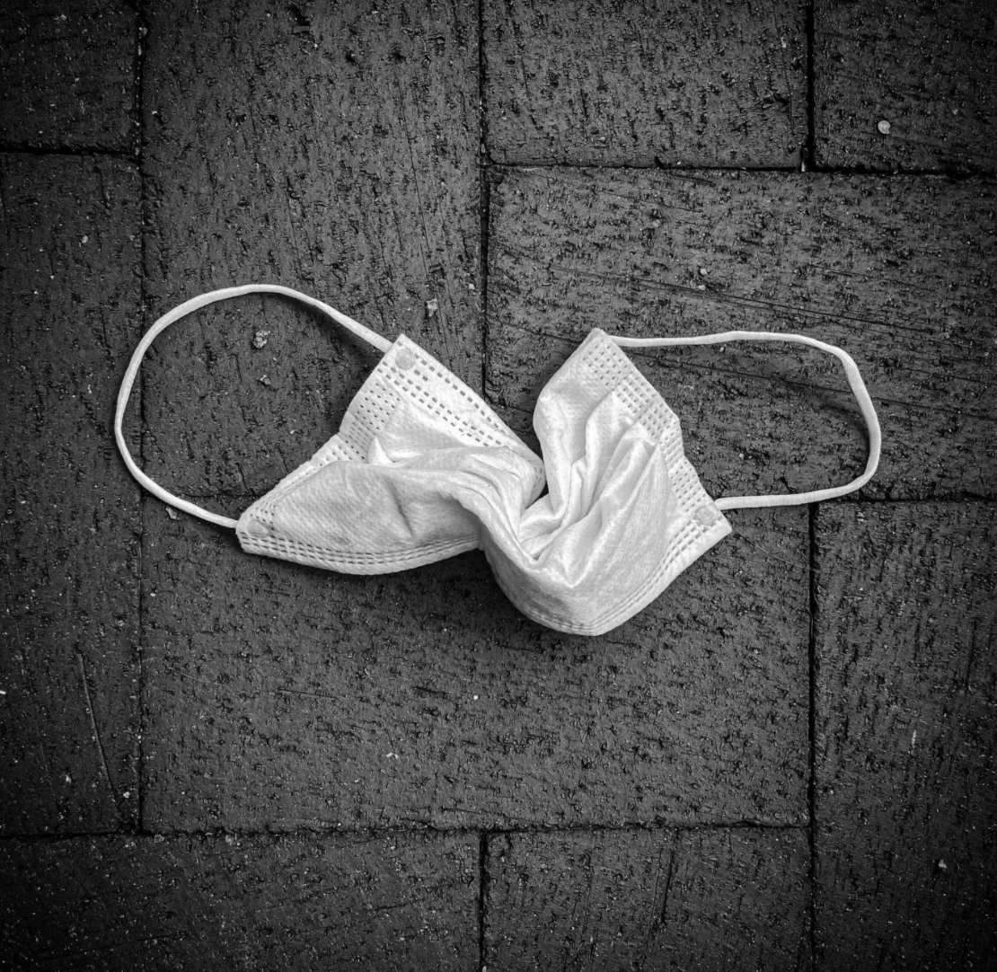  A black and white digital photograph by Jess Walters. A white surgical mask is lying on a brick sidewalk; it is crinkled in the middle with the ear loops laying out to either side. 